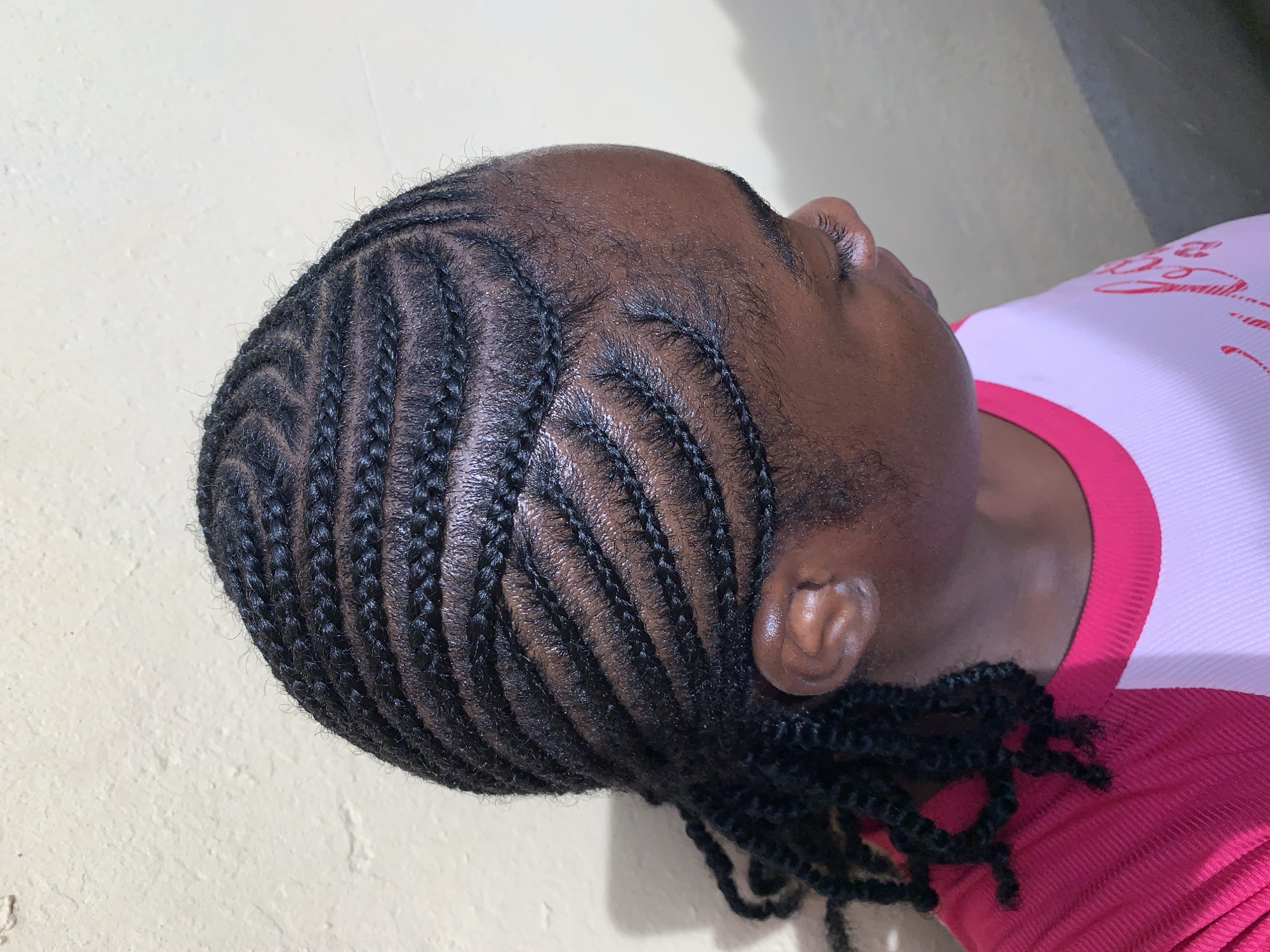 A close-up shot features an individual with cornrow braids. The braids are neatly arranged and dark in color. The person is wearing a shirt with a white body and a red collar and sleeves. The background is a light beige wall. The image does not provide information regarding the location in the city of Rafin Par, Nigeria.