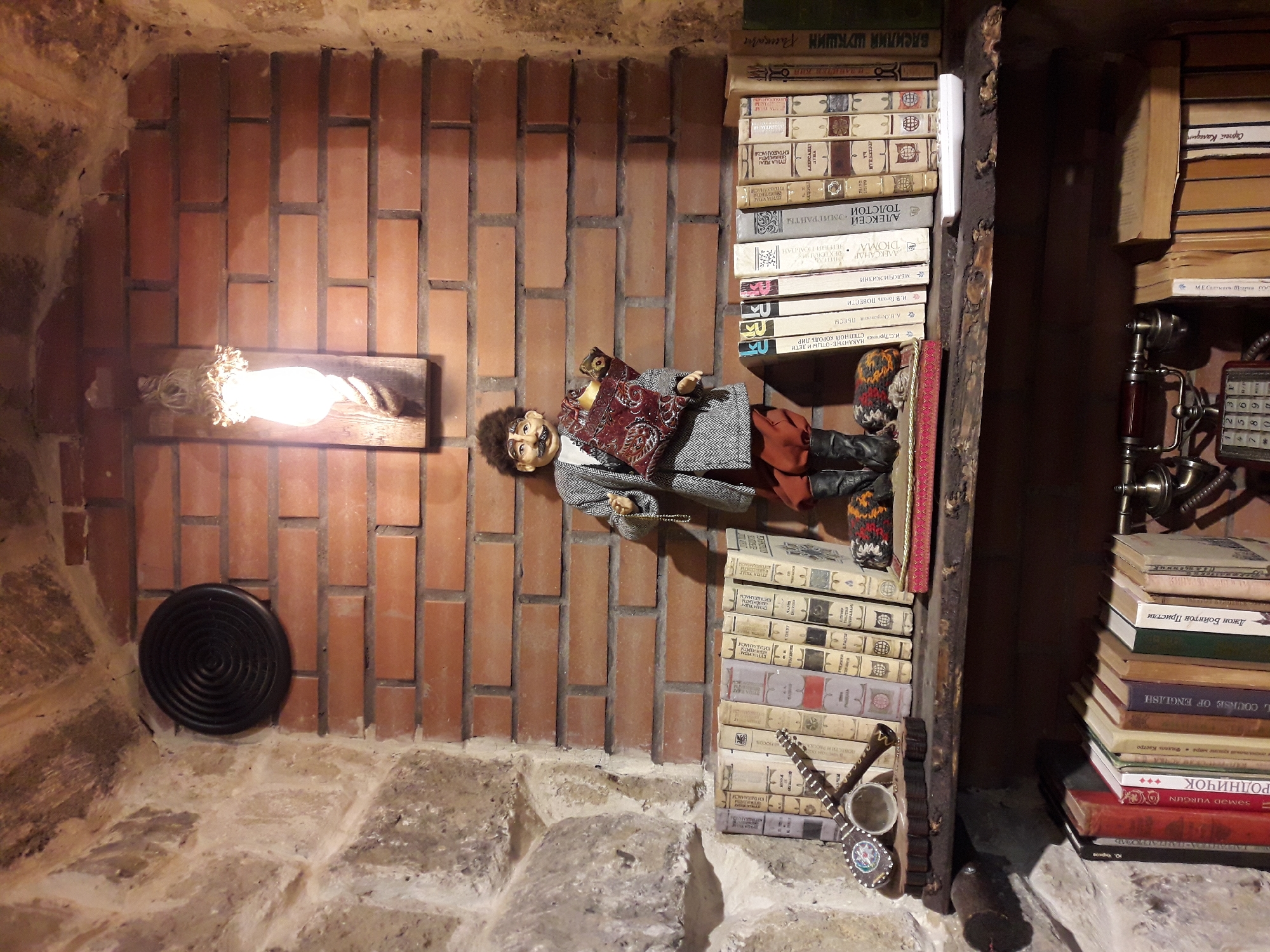 The image depicts a shelf built into a brick and stone wall. A doll, dressed in a grey and white patterned coat, orange pants, and black boots, stands on the shelf holding a book. The doll has dark curly hair and a mustache. To the left of the doll, a row of books is arranged, with a musical instrument resembling a saz or lute placed in front of them. Above the shelf, a light fixture with a rope and an Edison-style bulb hangs from the brick wall. To the left of the light, a black circular vent is mounted on the brick. Below the shelf, another row of books is visible, along with an antique telephone. The overall environment appears to be an indoor setting, possibly a room decorated with vintage items.