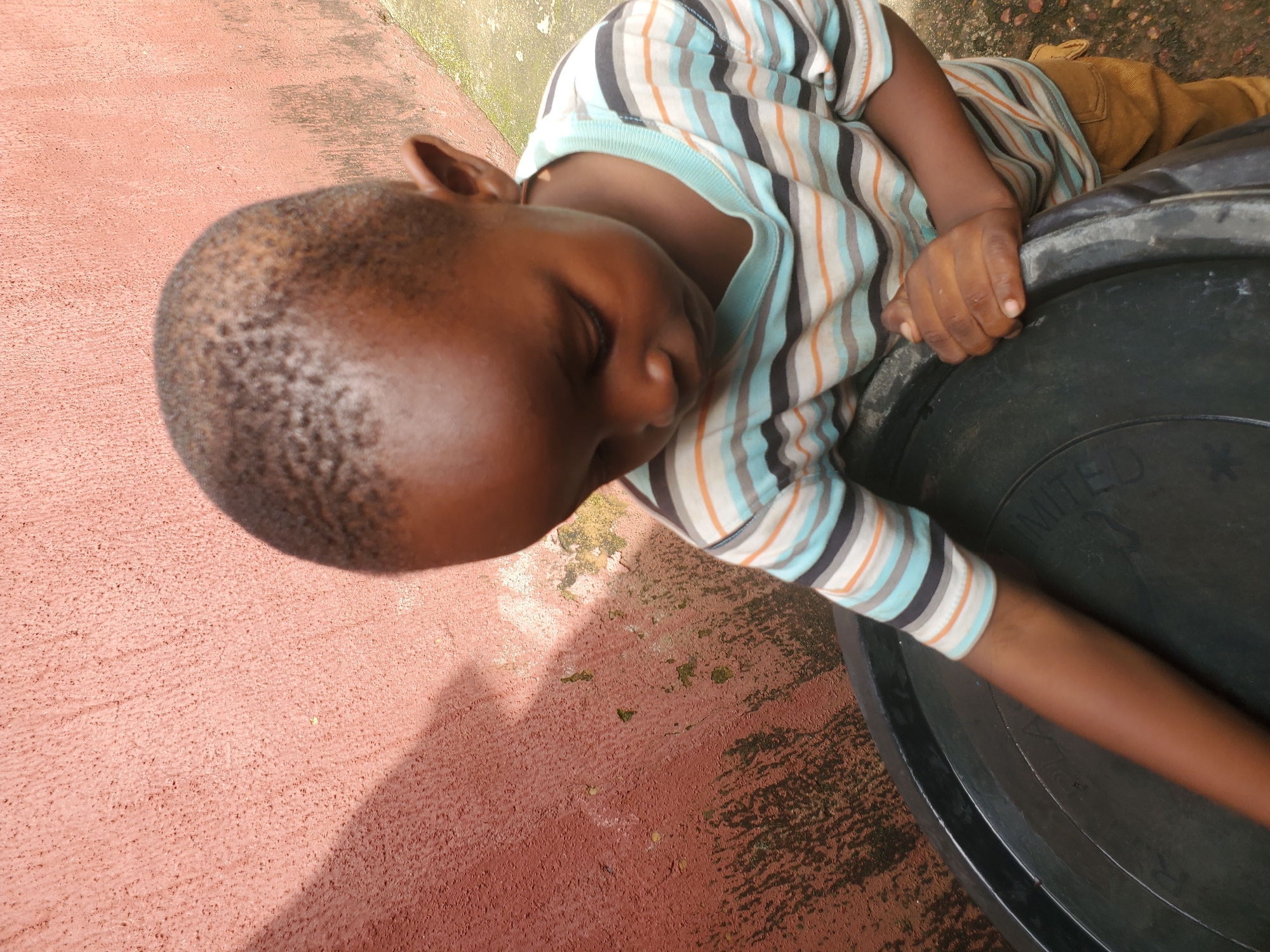 A young boy with dark skin and short hair is lying down, with his eyes closed, on a rough, reddish-brown surface. He is wearing a striped t-shirt with blue, white, orange, and black stripes, and brown pants. His left arm is resting on the rim of a large, black, circular object, possibly a container or a tub. The boy's head is tilted to the side, and his face appears relaxed.
The surface he is lying on has a textured appearance and is partially covered with some debris and shadows, suggesting an outdoor setting. In the background, there is a lighter-colored, textured surface, possibly a wall or pavement, with some patches of green moss or growth. The lighting indicates it is daytime, with strong shadows cast on the reddish surface, suggesting the sun is high.
The scene appears to be a casual moment, possibly the boy resting or napping. There is no specific event or activity clearly depicted beyond the boy's repose. The overall impression is one of a simple, everyday moment in an outdoor environment. Some faint text is visible on the black object, appearing as "UNITED" and part of another word. The location context of Jos, Nigeria, provides a cultural and geographical backdrop to the image.