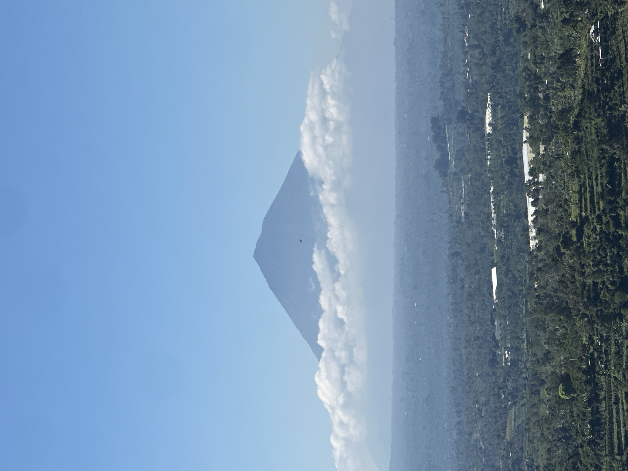 The image depicts a panoramic view of a mountainous landscape with a prominent volcano in the background, shrouded in clouds. The foreground shows a lush, green valley with terraced fields and scattered buildings, characteristic of the rural outskirts of Baturiti, Indonesia. The sky is clear and blue, with a single bird visible in flight near the volcano. The overall scene suggests a peaceful and scenic environment, likely captured on a clear day.