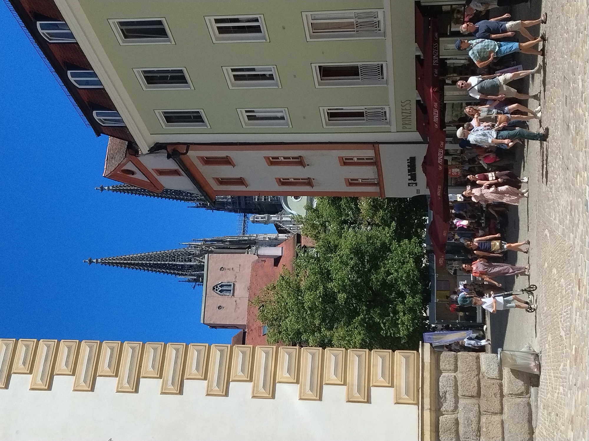 The image depicts a street scene in Regensburg, Germany, likely near the Regensburg Cathedral, identifiable by the prominent spires in the background. The foreground features a cobblestone street with several people walking and interacting.
On the left, a textured stone wall and a cream-colored building with decorative horizontal bands are visible. The street is populated with individuals dressed in casual summer attire. A young boy on a scooter is a focal point in the mid-ground, accompanied by two women. To the right, a light green building with multiple windows and dormer roofs dominates the scene. Below this building, there are awnings with signage that reads "PRINZESS Cafe" and "PRINZESS". People are seated at tables under these awnings, suggesting an outdoor cafe or restaurant area.
In the background, the ornate spires of a large Gothic cathedral rise against a clear blue sky, confirming the location in Regensburg. Trees provide shade in the mid-ground between the buildings and the cathedral. The overall atmosphere is that of a sunny day in a bustling European city square.