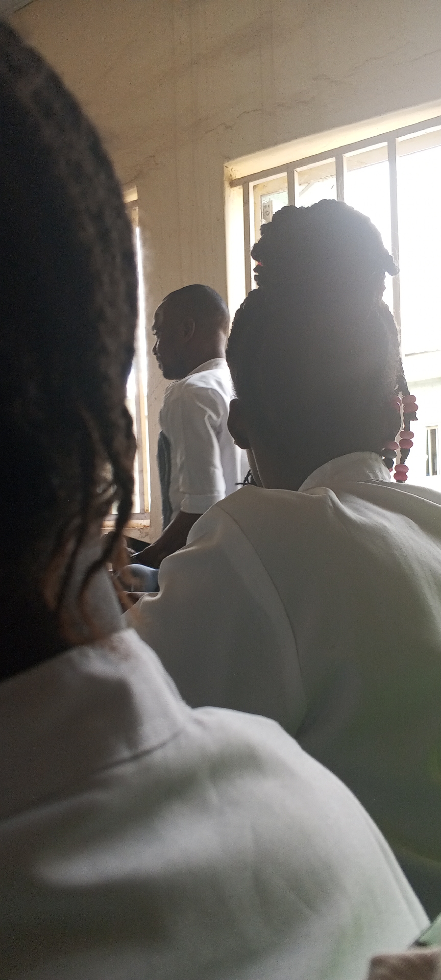 Location: Keffi, Nigeria
Description: The photograph depicts three individuals in an indoor setting, likely a classroom or meeting room. The foreground is dominated by the back of a person with dark, braided hair. To the right, another person with similarly styled hair is visible, adorned with beads. A man in a white shirt is positioned behind them, facing a window. The window provides natural light, illuminating the scene. The walls are light-colored. All three are presumed to be seated or standing in close proximity.