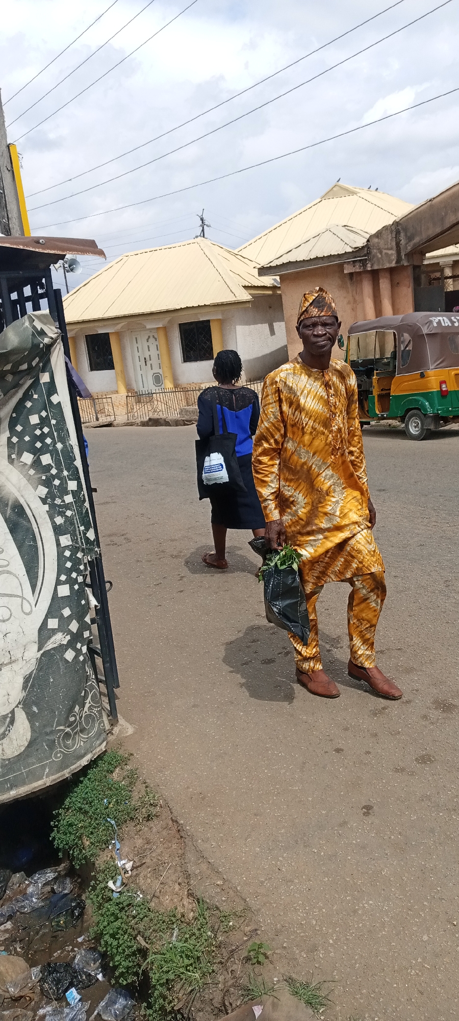 An elderly man in a traditional patterned yellow and brown outfit and matching headwear is walking towards the camera, holding a black plastic bag with green contents. A woman in a blue top and black skirt walks behind him, carrying a black tote bag. In the background, there is a three-wheeled vehicle (Tuk-Tuk) and houses with yellow roofs. In the foreground, there is a trash-filled drainage area and part of a weathered structure with graffiti. The location is identified as Jos, Nigeria, based on the presence of the Tuk-Tuk, common in the city.