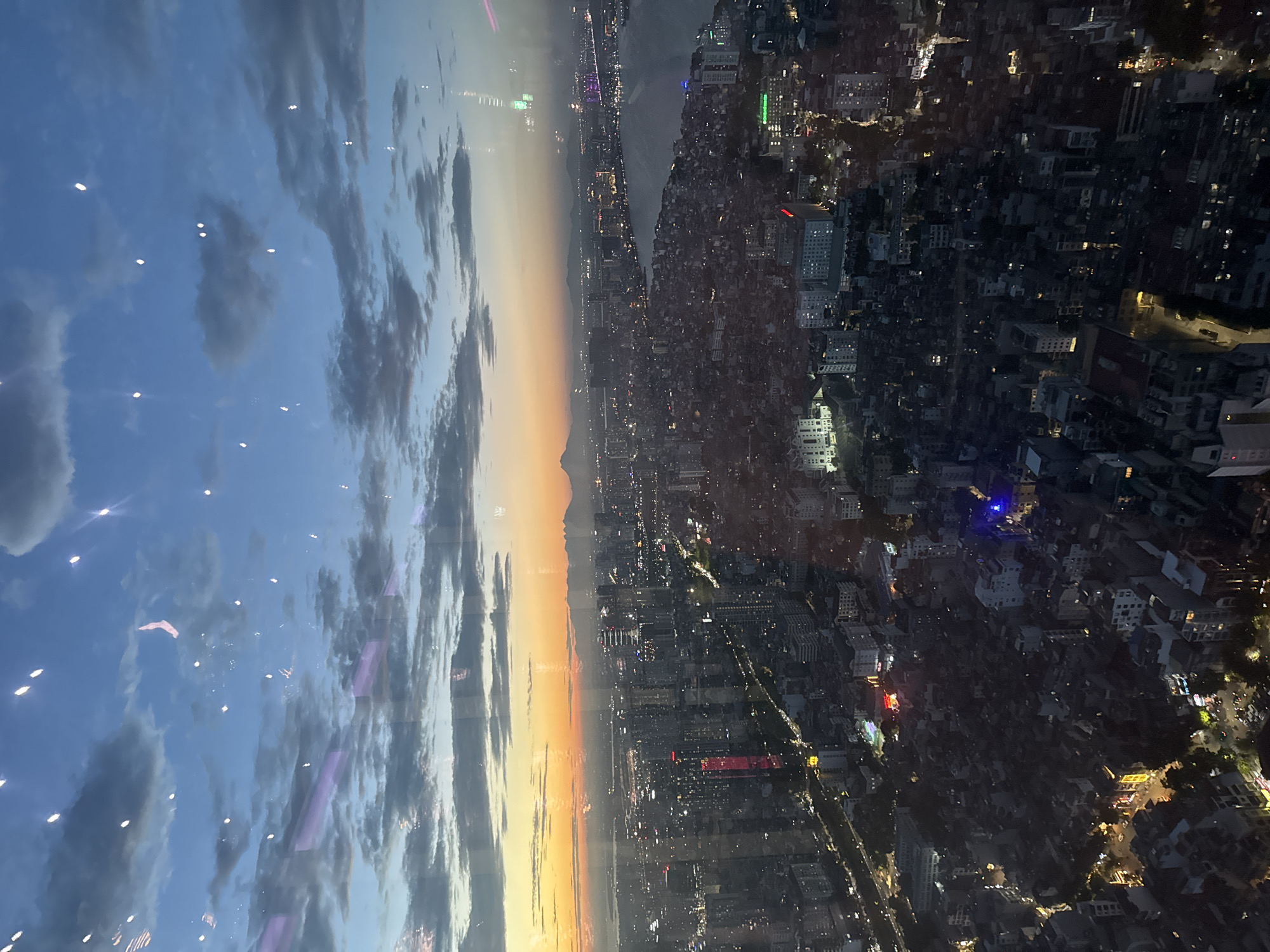 The image captures a sprawling cityscape at dusk, likely Hanoi, Vietnam, viewed from a high vantage point. The scene is divided vertically. The left side is dominated by a dramatic sky, transitioning from a deep blue with scattered clouds to a warm gradient of orange and yellow near the horizon, indicating sunset or twilight. Small, bright lights, possibly reflections or distant aircraft, are scattered across the sky.
The right side of the image showcases the urban landscape as darkness descends. Numerous buildings, varying in size and architectural style, are densely packed together. The city is illuminated by a myriad of lights emanating from windows, streetlights, and building facades, creating a vibrant, speckled pattern against the dark terrain. A body of water, likely a river or bay, is visible in the mid-ground, reflecting the city lights. Distant mountains can be seen silhouetted against the fading sky. The overall atmosphere is one of urban activity and the beauty of a city transitioning from day to night. No people are distinctly visible. The activity is primarily the unfolding of a natural phenomenon (sunset) and the continuous operation of a large city. There are no discernible text elements in the image.