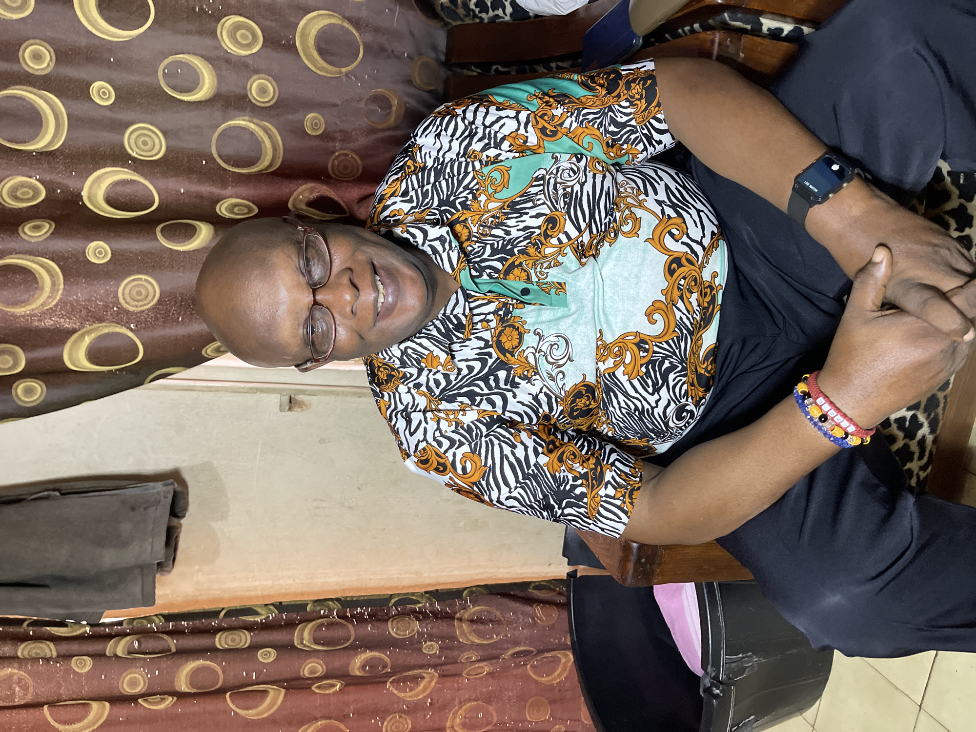 A Black man, wearing glasses, a patterned short-sleeved shirt, and black pants, sits in a wooden chair. He has a bald head and is smiling. He is wearing bracelets on his left wrist and a smartwatch on his right wrist. The background shows a patterned curtain and a wall. A black trash bin is visible to the left of the man. The scene appears to be indoors, in a residential setting, possibly in Jos, Nigeria.
