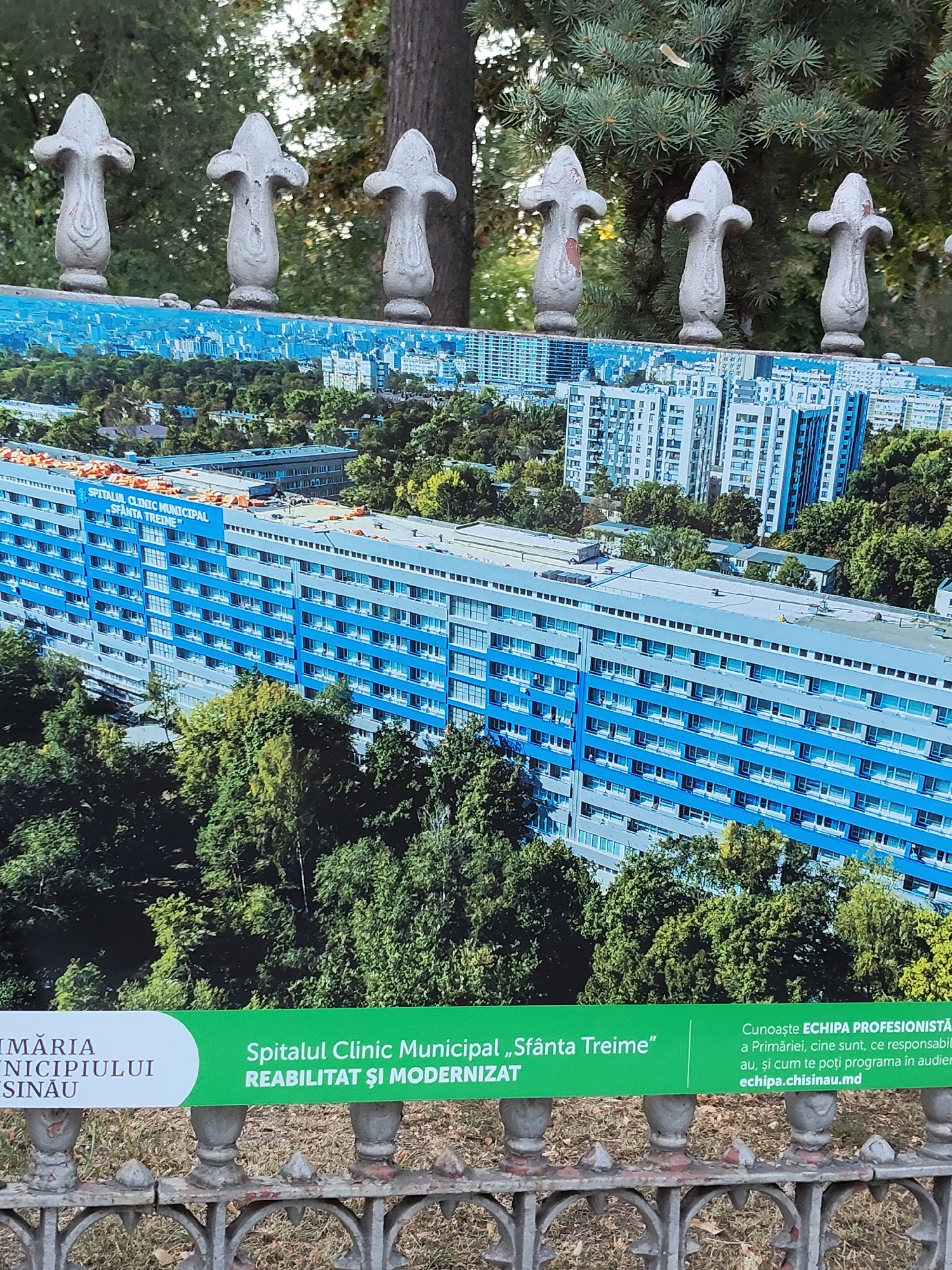 The image shows a photograph of a large, blue and gray hospital building. The building is several stories tall and appears to be modern. The photograph is displayed on a fence in an outdoor setting. The fence is made of metal and has decorative posts. Behind the fence and the photograph, there are trees and foliage. In the foreground, there is a green and white sign with text. The text on the sign is in Romanian and mentions "Spitalul Clinic Municipal Sfânta Treime" which translates to "Municipal Clinical Hospital Holy Trinity". It also mentions "Primăria Municipiului Chișinău" which means "Chisinau Municipality City Hall". The sign also includes a web address "echipa.chisinau.md". This indicates that the photograph is of the Municipal Clinical Hospital Holy Trinity in Chisinau, Moldova.