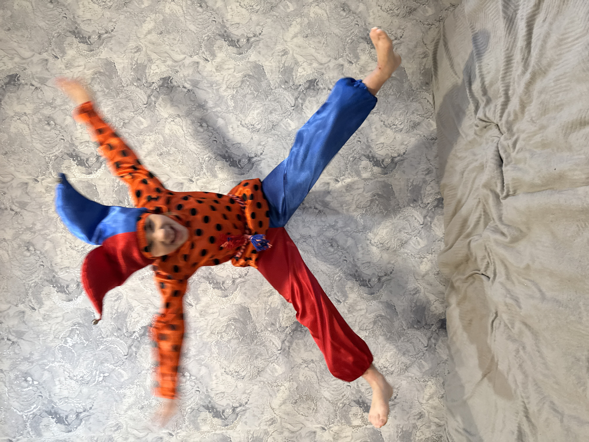 An indoor, low-angle photograph captures a child in mid-air, likely jumping or performing an acrobatic move, dressed in a vibrant, mismatched harlequin-style costume. The child's body is splayed out, forming an "X" shape, with bare feet extended. The costume features an orange top covered in black polka dots, one blue sleeve, one orange polka-dotted sleeve, and mismatched pant legs – one red and one blue. A pointed hat, split between red and blue, is worn on the child's head. A wide, joyful smile is visible on the child's face, indicating playfulness and happiness, despite significant motion blur evident across their moving limbs and body.
The background features a patterned wall covered in abstract gray and white swirling wallpaper. To the right of the frame, a textured, light-colored blanket or duvet, possibly on a bed, is partially visible, suggesting a bedroom or living space. The lighting is diffused and appears consistent with an indoor setting. There is no visible text. The overall scene captures a moment of energetic, lighthearted play. The context of the location is Rusanovo, Russia.