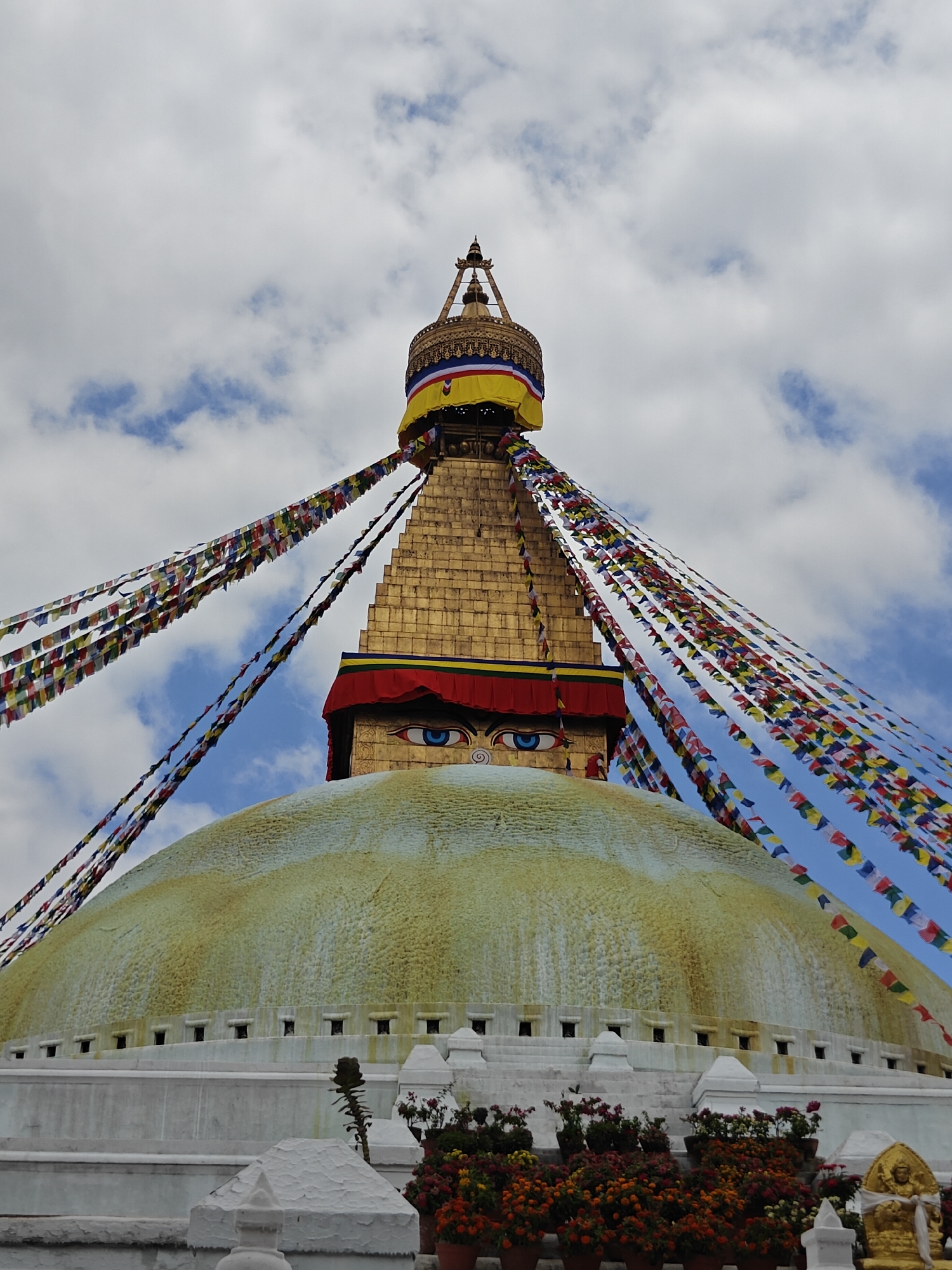 This image shows the Boudhanath Stupa in Gokarneshwar, Nepal, a UNESCO World Heritage site. The stupa is a large dome with a golden spire topped with a canopy. Prayer flags in various colors are strung from the spire, fluttering in the sky. The eyes of Buddha are painted on the sides of the spire. The base of the stupa is a white circular structure with multiple levels. In the foreground, potted plants with colorful flowers are arranged. The sky above is cloudy with patches of blue.