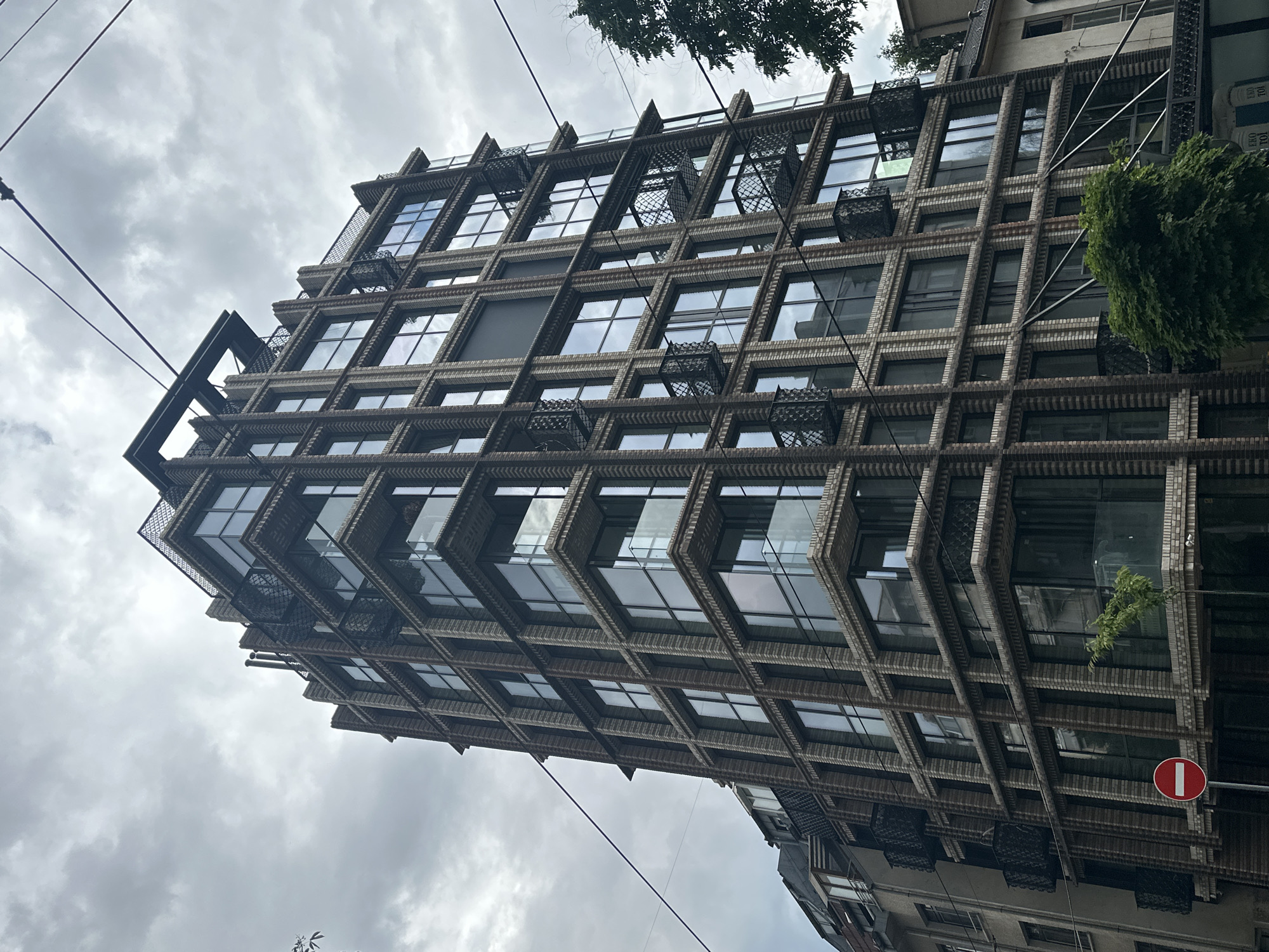 A low-angle perspective image shows a tall, modern building in Sofia, Bulgaria, under a cloudy sky. The building has a distinctive facade with a grid-like pattern of brick or stone framing large windows. Small, decorative wrought-iron balconies are attached to some of the windows. Electrical wires crisscross the sky above the building. A "no entry" sign (white horizontal bar on a red circle) is visible on the lower right. The building is partially obscured by tree foliage. Adjacent buildings are partially visible at the bottom edge.