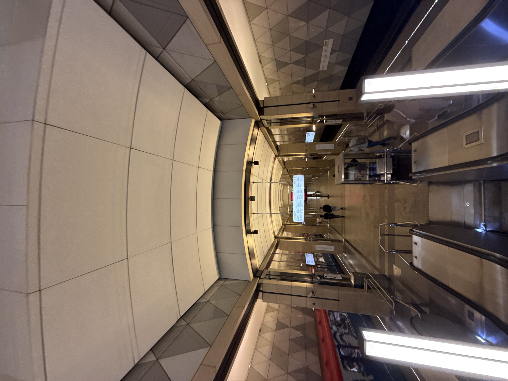 The image depicts the interior of a modern metro station in Moscow, Russia. The station features a high, arched ceiling made of light-colored panels with subtle geometric patterns. The walls are adorned with a repeating triangular motif in shades of gray. On the left side of the platform, a train with a red stripe is visible, its windows reflecting the station lights.
In the foreground, two escalators are prominent, each illuminated by tall, rectangular vertical lights. The escalators are made of polished metal.
Further down the platform, several people are walking. Two individuals are seen walking away from the camera, and another person is walking towards the camera, carrying a backpack. A few other figures are visible in the distance, some near a digital display board mounted overhead. The floor of the platform is tiled with a neutral color. The overall impression is one of clean lines, modern design, and efficient public transportation.