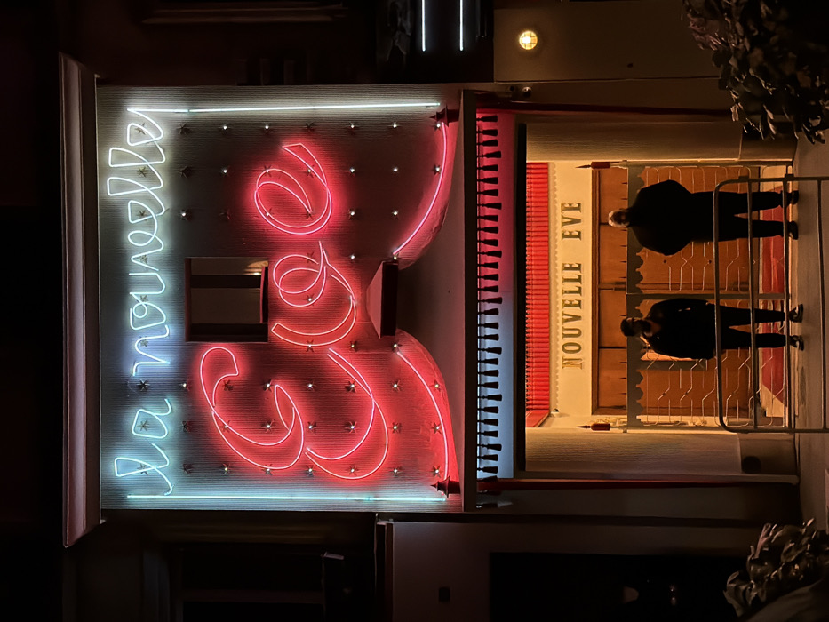The La Nouvelle Eve cabaret entrance at night features a vibrant neon sign, with the venue's name in illuminated lettering. Two security guards stand in front of the entrance, which is protected by a gated barrier. The building is illuminated, with additional lights above the neon sign.