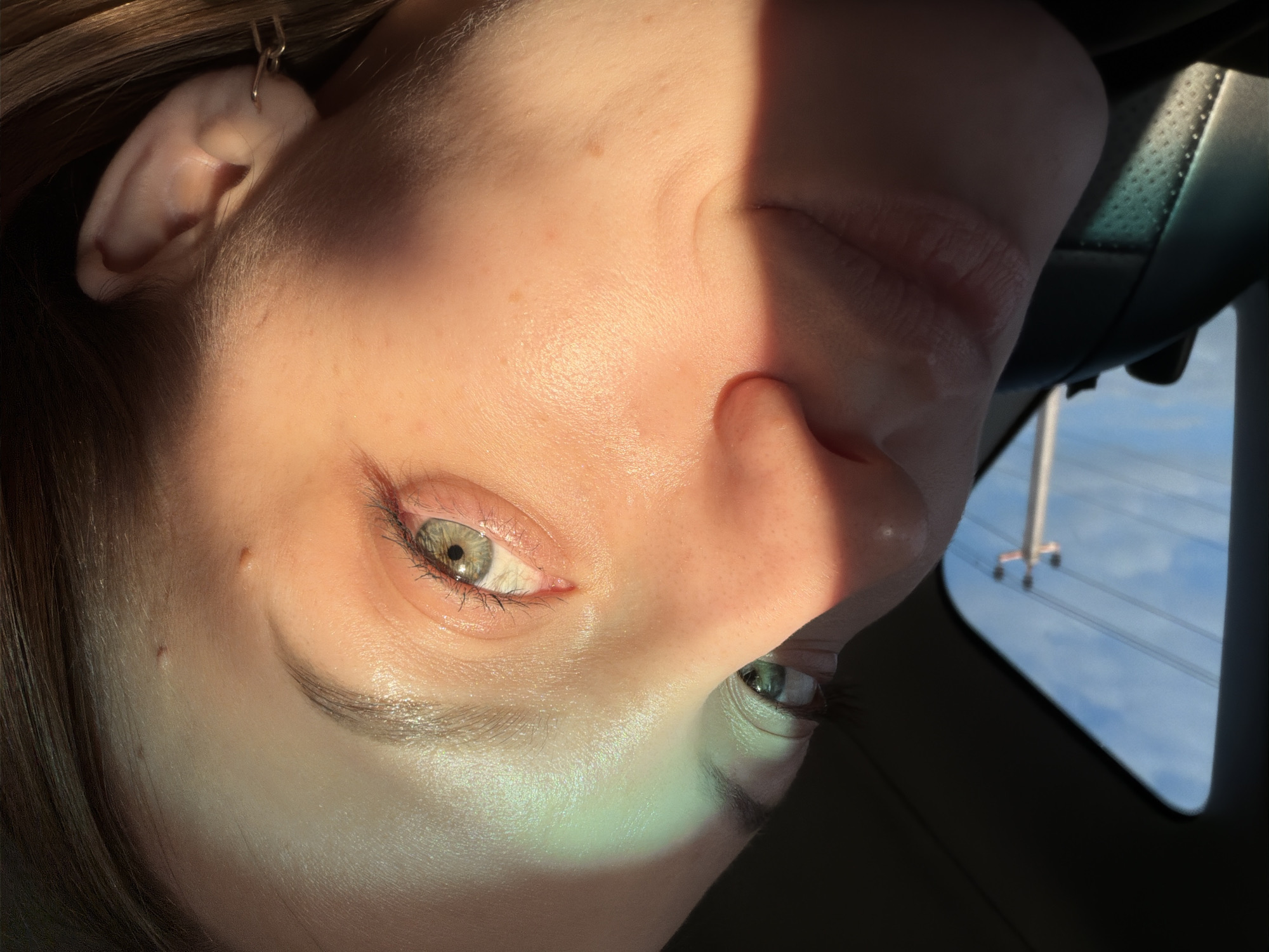 A close-up, rotated view captures the face of a light-skinned individual, likely female, with green eyes. The face is partially illuminated by sunlight, with shadows bisecting the face vertically. Details visible include freckles, eyelashes, and an earring. In the background, seen through what appears to be a car window, there are utility poles against a blue sky. This location is not recognizably in Minino, Russia, though the utility poles are consistent with Russian infrastructure.