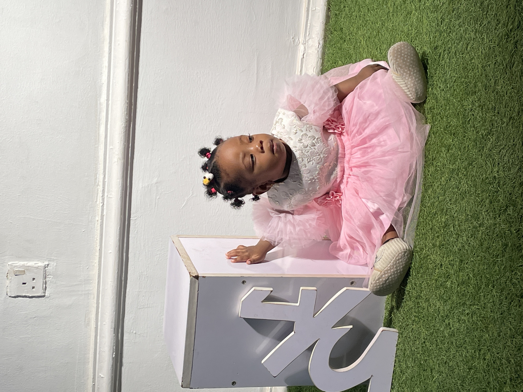 A young child, likely a toddler, is captured in a full-body shot, seated on an artificial green grass surface. The child is dressed in a white top with an embossed floral pattern and a voluminous light pink tutu-style skirt featuring ruffled sleeves and decorative pink flowers. Their dark hair is styled into several small puffs, adorned with colorful hair ties, including one shaped like a yellow duck or chick. Light-colored, soft-soled shoes cover their feet.
The child is positioned with their left hand resting on a white, rectangular box or pedestal. Their head is tilted slightly upwards, and they gaze towards the viewer's left with a neutral or thoughtful expression. Behind the child and to their left is a white wall with a prominent vertical decorative molding. A white Type G electrical outlet, commonly used in Nigeria, is visible on this wall. Partial light grey letters, resembling "KG," are visible on the side of the white box.
The scene suggests an indoor setting, likely a studio or a specially decorated area for a photoshoot, possibly celebrating a special occasion. The power outlet serves as a location cue, consistent with the provided context of Barakin Akawo, Nigeria.