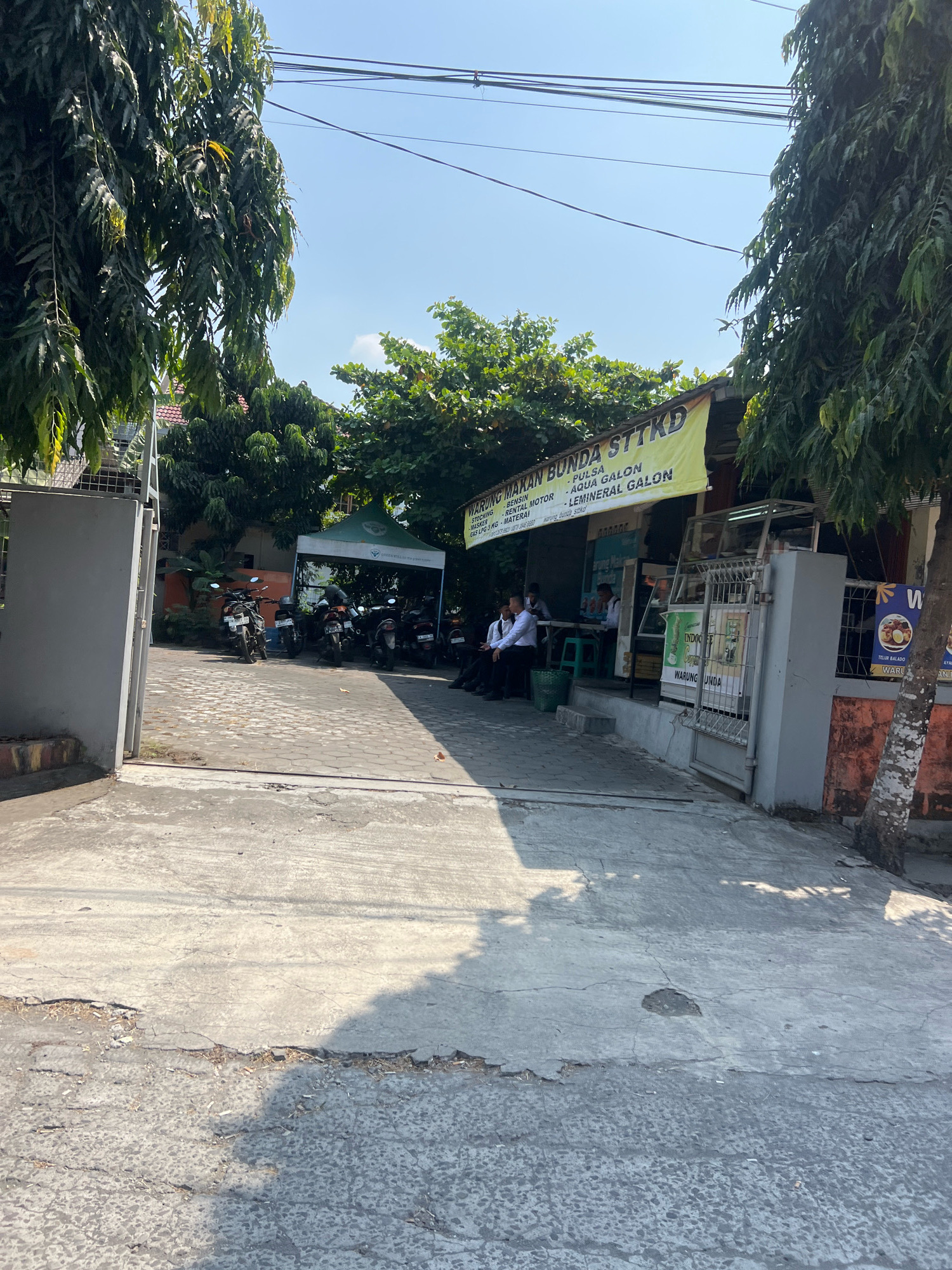 The image depicts the exterior of a warung (small eatery) in Sewon, Indonesia, under a clear blue sky. A large, faded yellow banner hangs above the warung, advertising "MAKAN BUNGA STTKD" and listing various items like "PULSA," "AQUA GALON," and "LEMINERAL GALON."
In front of the warung, several motorcycles are parked. People, some appearing to be employees wearing white shirts, are visible behind a counter. Others are seated on stools. A green tent is set up to the left of the warung entrance.
The ground in front of the warung is paved with concrete and cobbles, showing signs of wear and tear with cracks and patches. To the left, a gray metal gate stands open, leading into the premises. Lush green trees with long, drooping leaves partially obscure the view of the buildings. To the right, a tree with textured bark grows alongside the warung. A poster featuring food items is visible on the right side of the warung. The overall scene suggests a busy, casual establishment in a tropical setting.