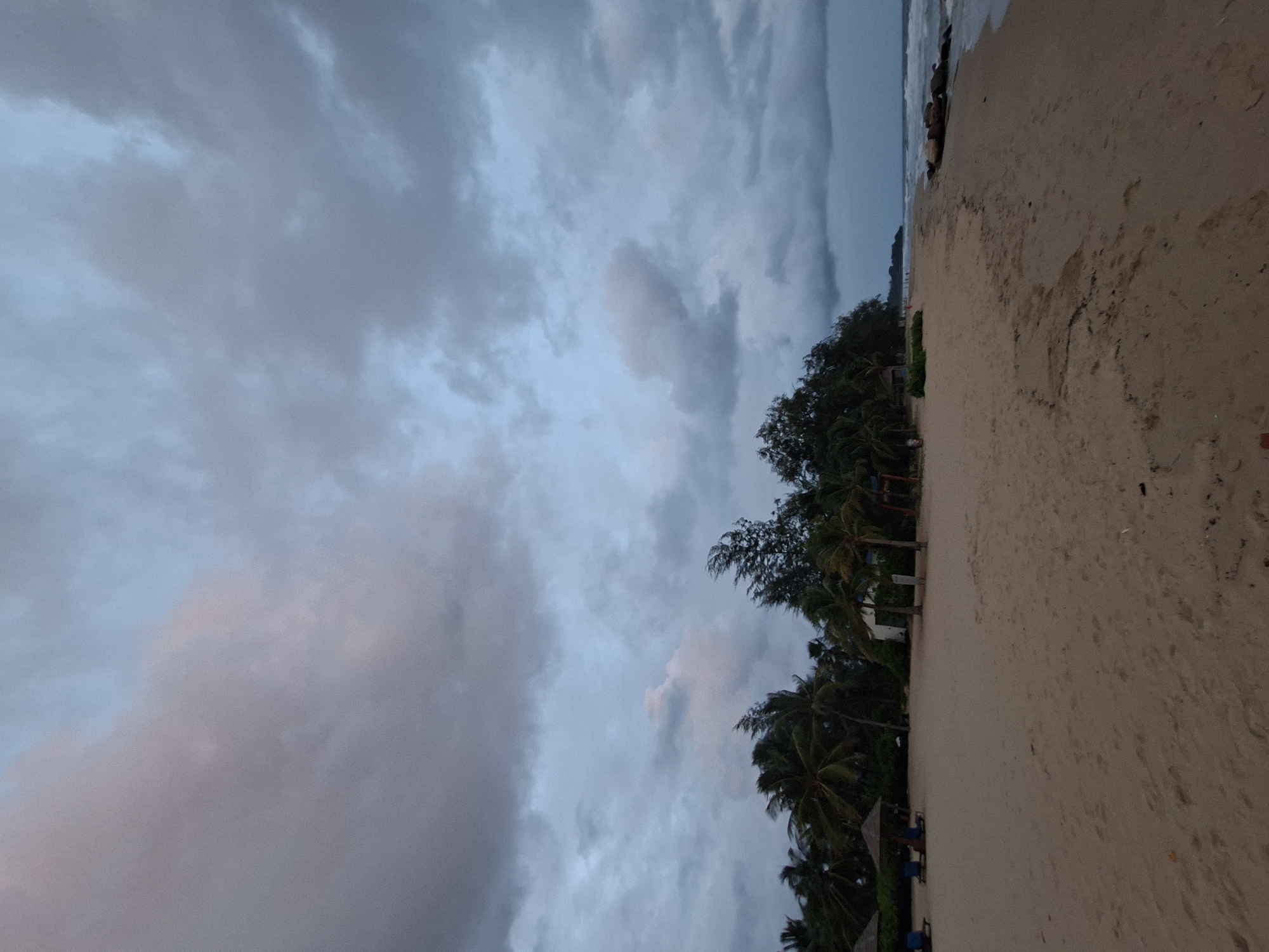 This image captures a serene beach scene on the island of Phú Quốc, Vietnam. The foreground is dominated by a stretch of pale sand, with gentle waves lapping at the shore on the right. In the mid-ground, several palm trees and other lush vegetation line the edge of the beach, interspersed with what appear to be small huts or structures. Beach umbrellas and lounge chairs are visible on the left, suggesting a resort or a public beach area. The sky above is filled with dramatic, dark gray clouds, hinting at an impending storm or the aftermath of one. The overall atmosphere is moody and tranquil, with the natural beauty of the tropical landscape contrasting with the foreboding sky.