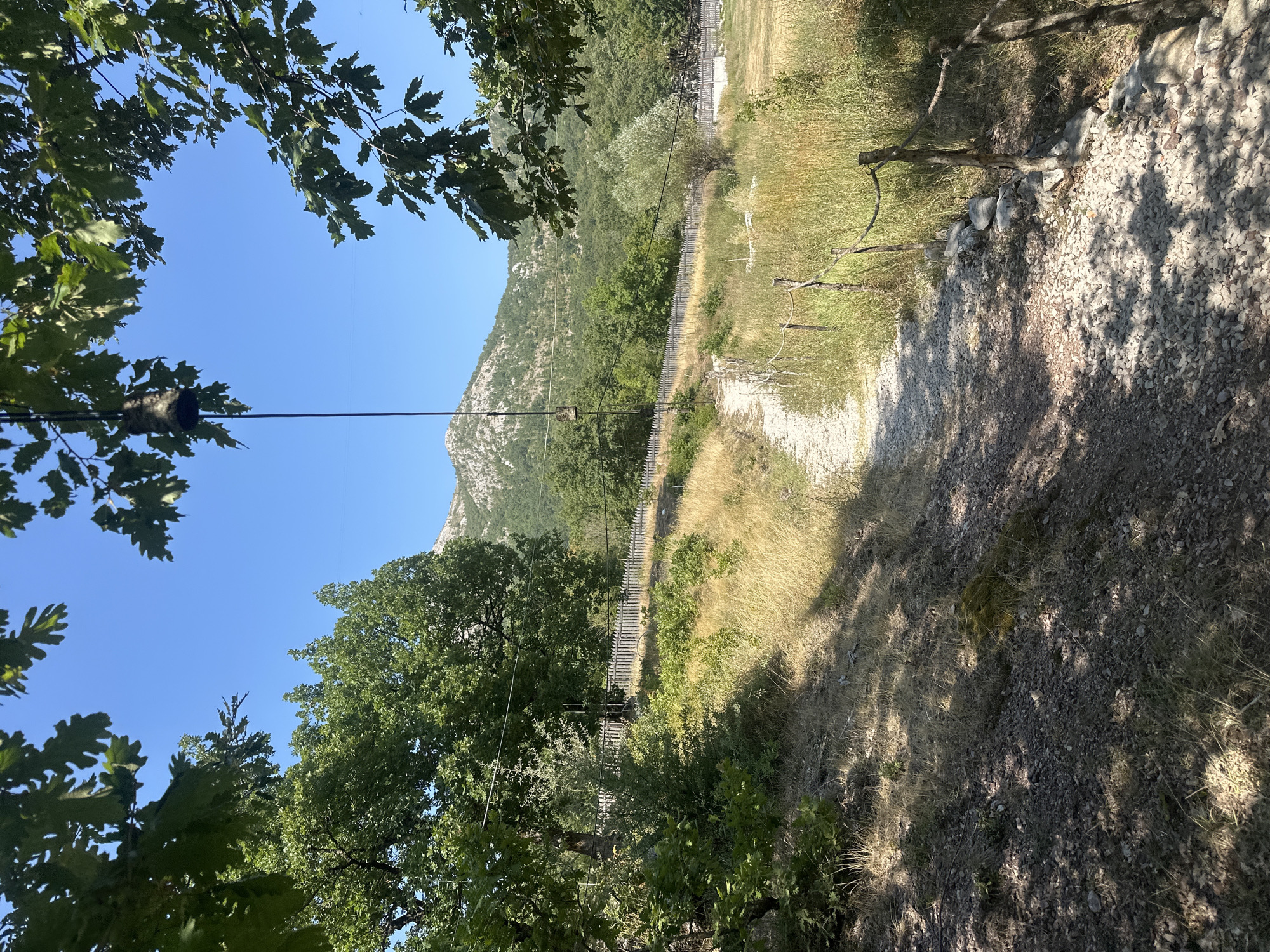 The image depicts a path winding through a rural, mountainous landscape under a clear blue sky. The path is composed of gravel and stones and is bordered by dry, tall grass and a simple wooden fence. Trees, primarily with large, lobed leaves, frame the scene from above and to the sides. In the background, a gentle slope rises to form a mountain range covered in dense green foliage and rocky outcrops. A single, dark pole with some attached equipment stands near the path, and a zipline cable is visible stretching across the upper part of the image, suspended from a tree. The overall impression is one of a natural, untamed environment, possibly a hiking trail or recreational area. The scene is static, with no people or animals visible, and no apparent action taking place.