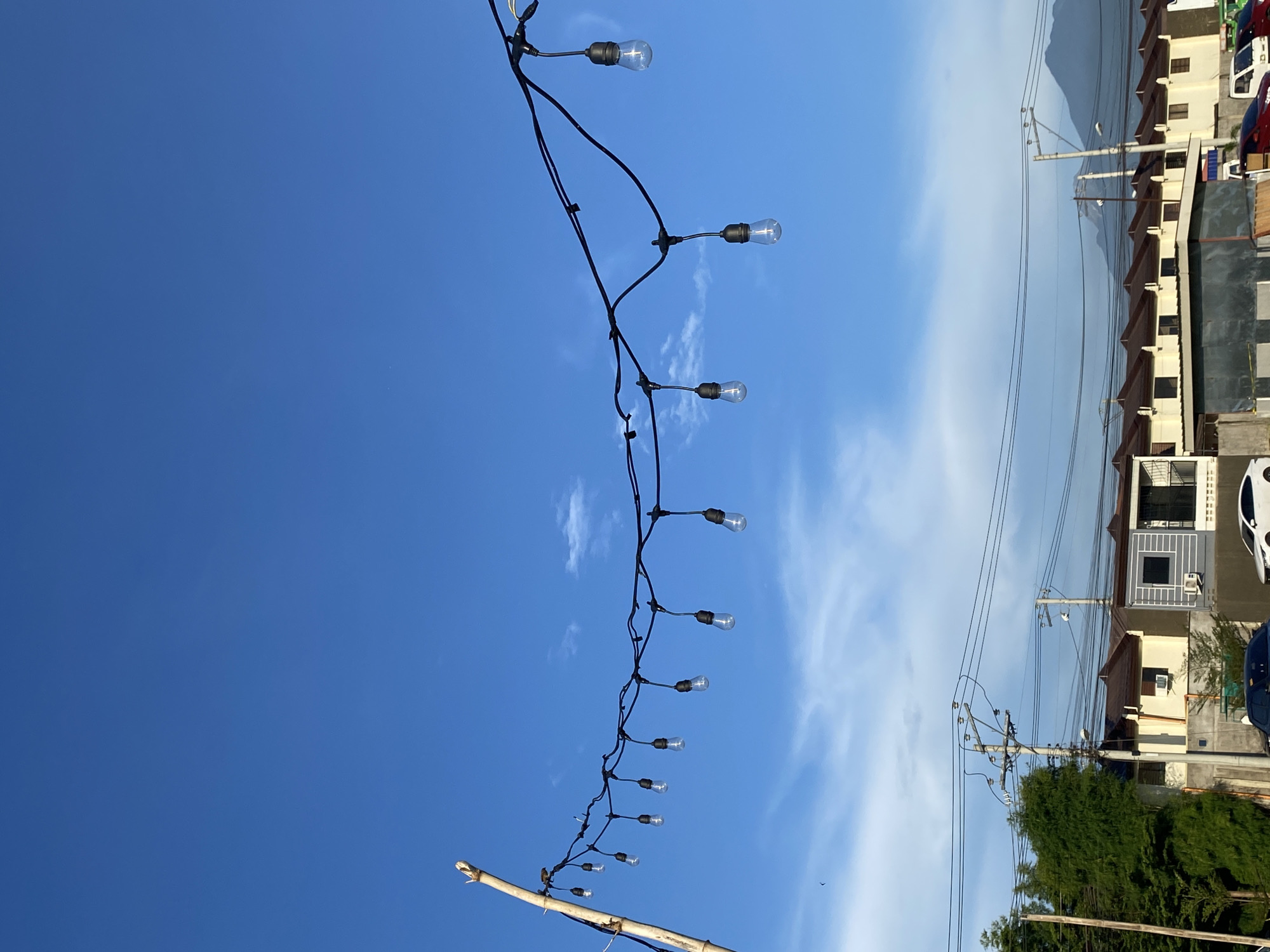 In the city of Davao City, Philippines, a string of clear Edison bulbs hangs suspended against a bright blue sky dotted with wispy clouds. The lights are attached to a black wire supported by a thin bamboo pole on the left and presumably anchored further to the right, out of frame. Below the string of lights, a row of houses with brown roofs stretches across the mid-ground, interspersed with tall green trees and utility poles carrying multiple wires. In the distance, the silhouette of a mountain range is visible against the horizon. Several cars are parked in front of the houses, suggesting a residential area. The overall scene depicts an outdoor setting, likely a street or residential compound, under a clear sky.