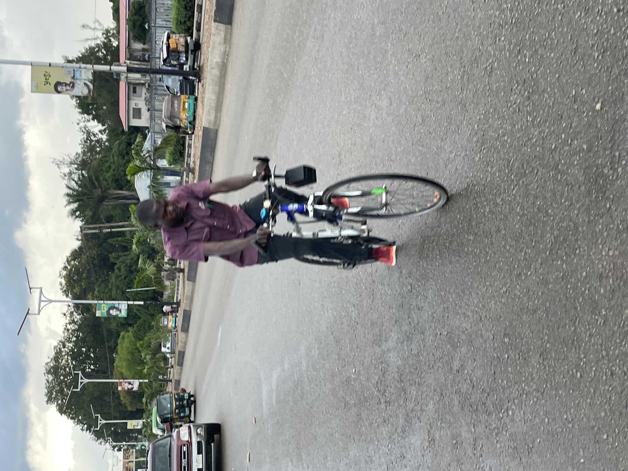 The image depicts a street scene in Jos, Nigeria, under an overcast sky. A man is riding a bicycle in the foreground, wearing a maroon polo shirt and dark pants. His bicycle is blue and white, with a black bag attached to the handlebars. Red and green reflectors are visible on the front wheel. The man's left foot is shod in a red athletic shoe.
The road is wet, suggesting recent rain, with visible tire tracks and patches of standing water. In the background, various vehicles are present, including a maroon SUV on the left, a green auto-rickshaw, and other vehicles further down the road. Billboards and streetlights line the side of the road, with lush green trees forming a backdrop. Buildings with red roofs are also visible in the distance. The overall atmosphere is that of a typical day on a busy street in Jos, Nigeria.