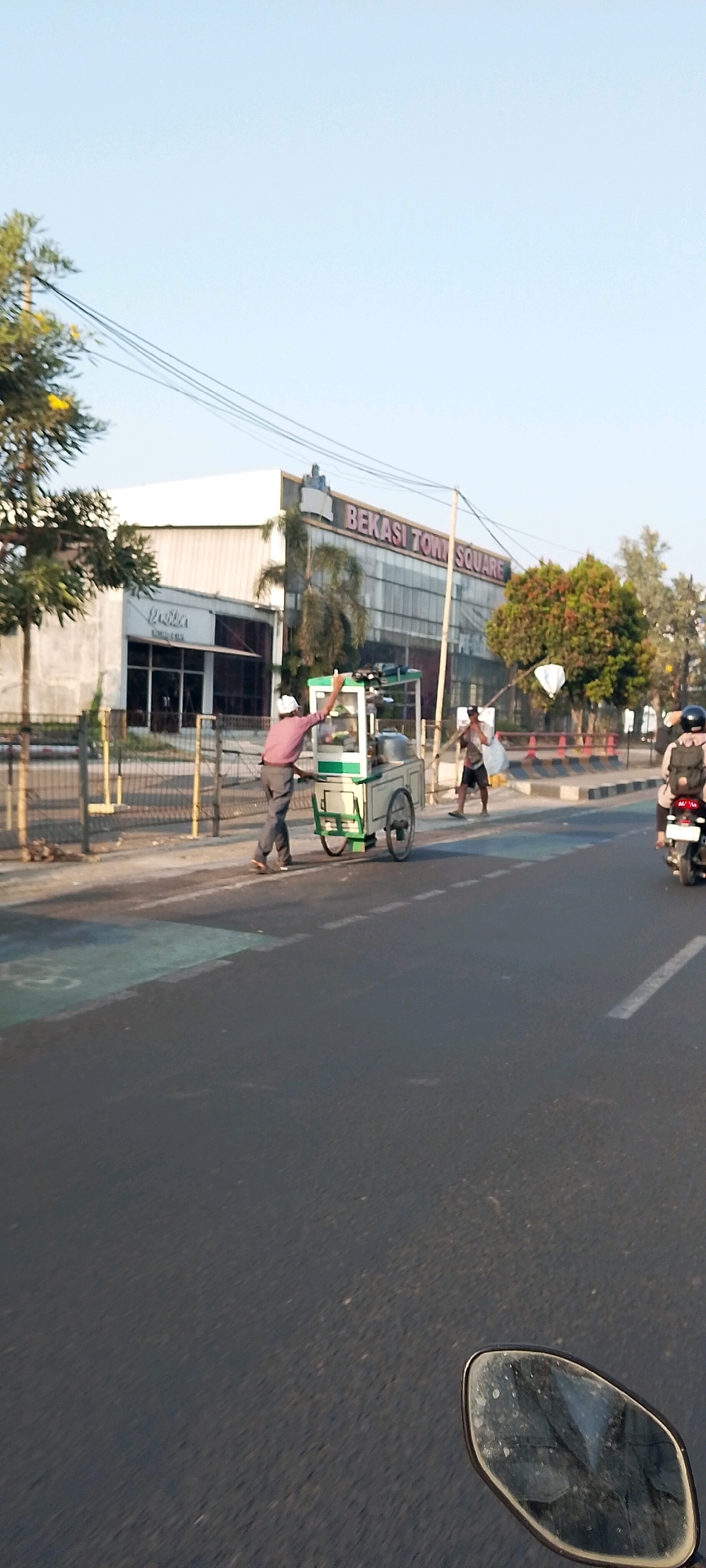 In Bekasi, Indonesia, a street scene unfolds with a focus on pedestrian activity and urban infrastructure. In the foreground, the asphalt road is marked with white dashed lines and a painted green section, possibly indicating a pedestrian crossing or bike lane. To the left, a metal fence borders a sidewalk area with sparse vegetation and yellow flowers on a tree. A man, wearing a pink shirt and gray pants, is pushing a green and white food cart with two wheels. He is captured mid-action, pushing the cart forward. Behind him, another man, wearing dark shorts and a light-colored shirt, is seen carrying a load on his shoulder. Further back, a motorcycle with a rider wearing a helmet and backpack is visible on the right side of the road, moving away from the viewer.
In the background, the "BEKASI TOWN SQUARE" building stands prominently, characterized by its modern architecture with large glass windows and a visible sign. To the left of the square, another building with a sign displaying "Dexter" is partially visible. The sky is clear and blue, suggesting fair weather. The scene depicts a bustling urban environment, with everyday activities of street vendors and commuters taking place.