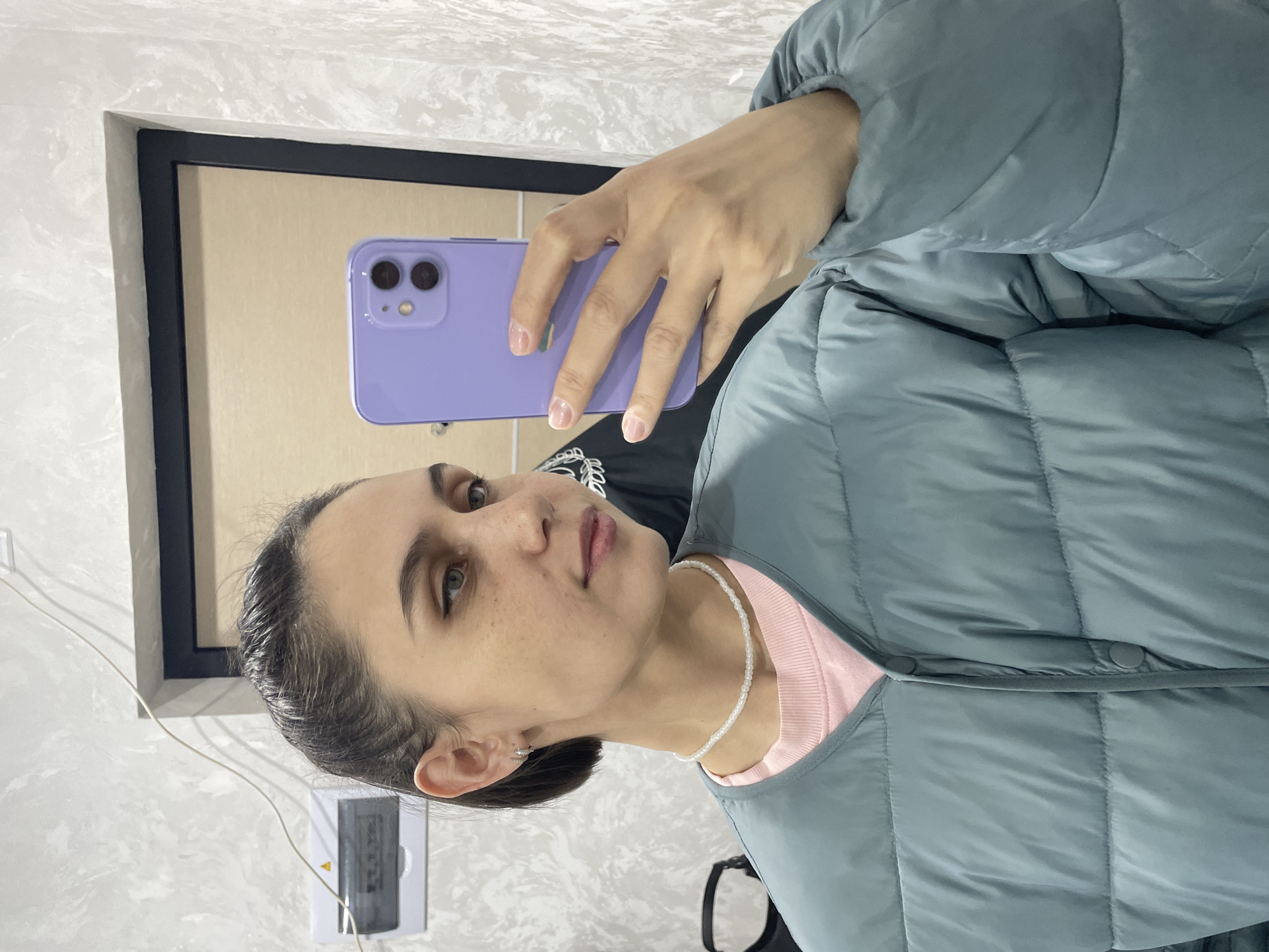 A woman is taking a selfie in a room. She is holding a purple smartphone in her right hand. She is wearing a light blue quilted jacket over a pink shirt and a white beaded necklace. The background includes a framed mirror or artwork and a white wall with an electrical outlet. The location is not discernible from the image provided.