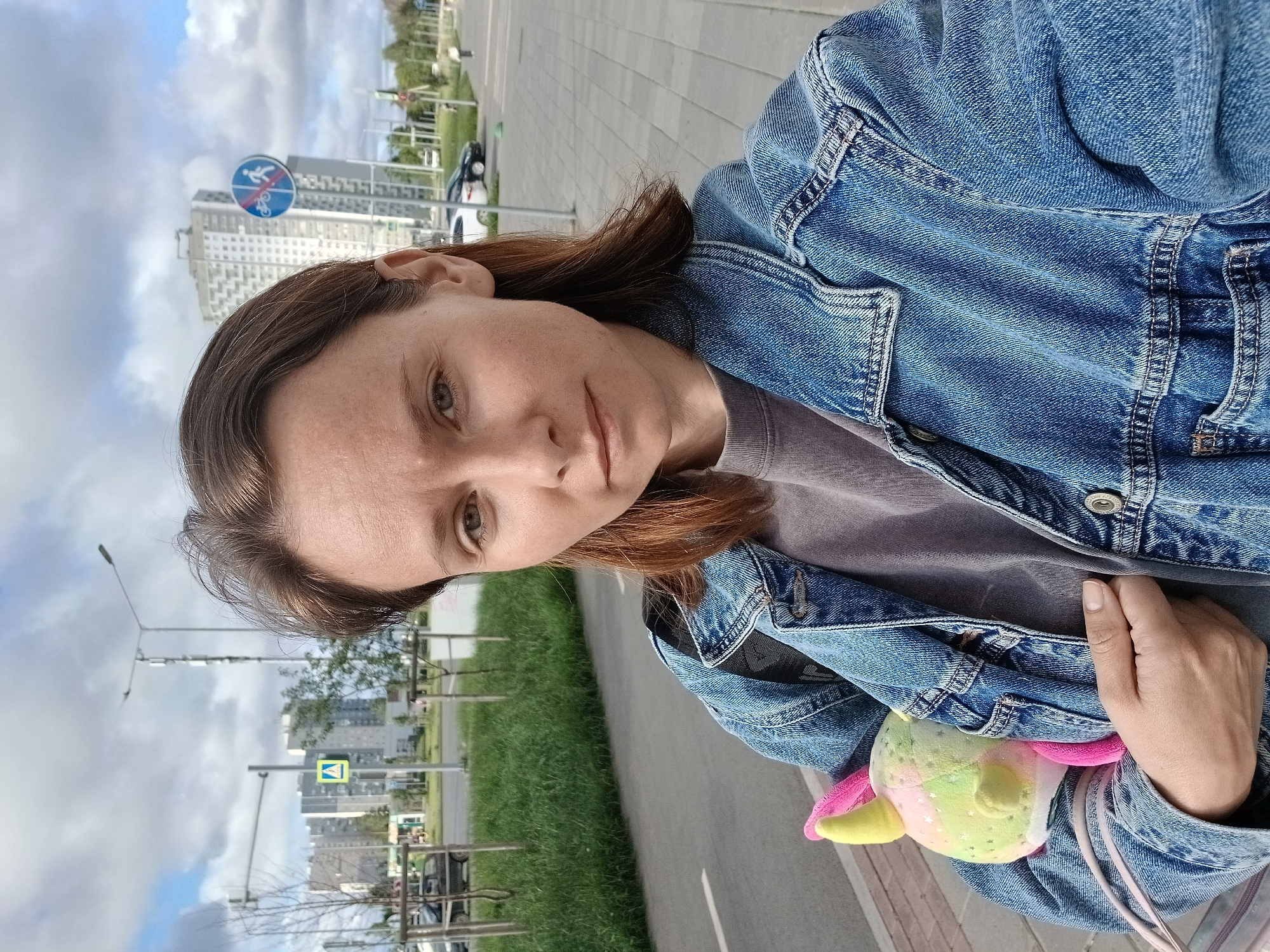 A woman with shoulder-length brown hair takes a selfie outdoors on a cloudy day in Yekaterinburg, Russia. She is wearing a denim jacket over a gray t-shirt and is holding a stuffed toy unicorn in her left arm. In the background, there is a sidewalk, a street with cars, a grassy area, and tall buildings. Traffic signs are visible, including a pedestrian crossing sign and a sign prohibiting bicycles and pedestrians. The woman is positioned in the foreground, with the city landscape behind her.