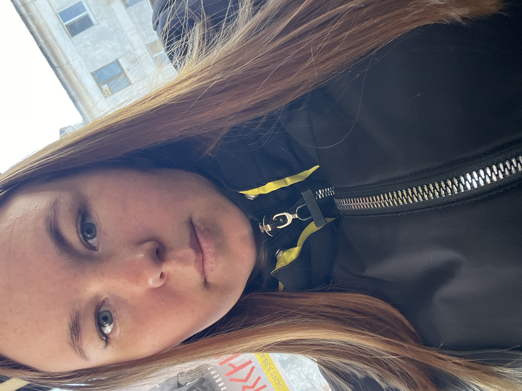 A selfie was taken in Chaikovsky, Russia, with a young woman as the primary subject. The image is oriented at an angle. The woman has fair skin, light eyes, and medium-length brown hair. She is wearing a black jacket with a silver zipper and yellow accents. The background includes a multi-story building with rectangular windows. A partially visible magazine with Russian text is also present.