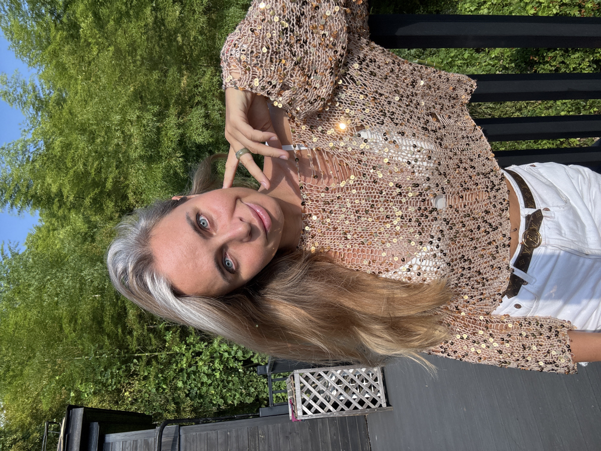 The image captures a woman posing outdoors against a backdrop of lush green bamboo trees and a clear blue sky. She is leaning on a black railing, with her right hand touching her cheek, adorned with a green ring. The woman has long, blonde hair with streaks of grey and striking blue eyes. She is wearing a cropped, open-knit sweater covered in gold sequins, revealing a white bra underneath. Below, she sports white denim shorts and a brown belt with a Louis Vuitton buckle. To her left, a dark wooden structure and a lattice-style wooden bin are visible. The setting suggests a sunny day in a natural environment, possibly a terrace or balcony. The specific location in Wuxing District, China, is not discernible from the visual information provided.