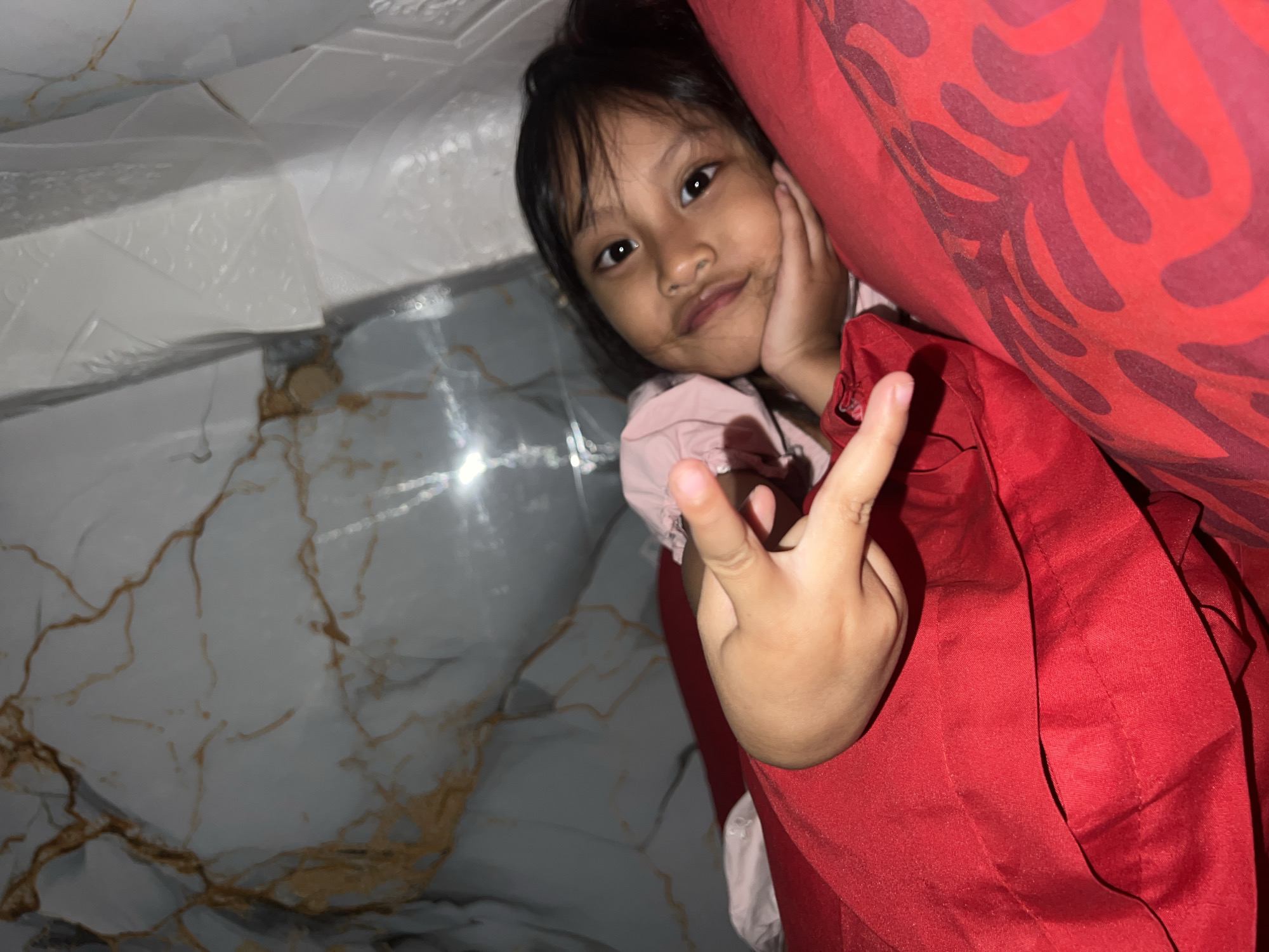 A young girl is lying down and looking at the camera. She is wearing a pink shirt and a red outer garment. Her right hand is resting on her cheek, and her left hand is held up with two fingers extended, as if making a peace sign. She has dark hair and brown eyes, and her expression is somewhat playful. She appears to be indoors, with a marbled floor visible in the background. A red fabric with a patterned design is draped over her, and another red garment is visible in the foreground. The lighting suggests it could be daytime, but the setting is not clear enough to determine the exact time or specific location beyond being indoors. The overall mood of the image is casual and lighthearted.