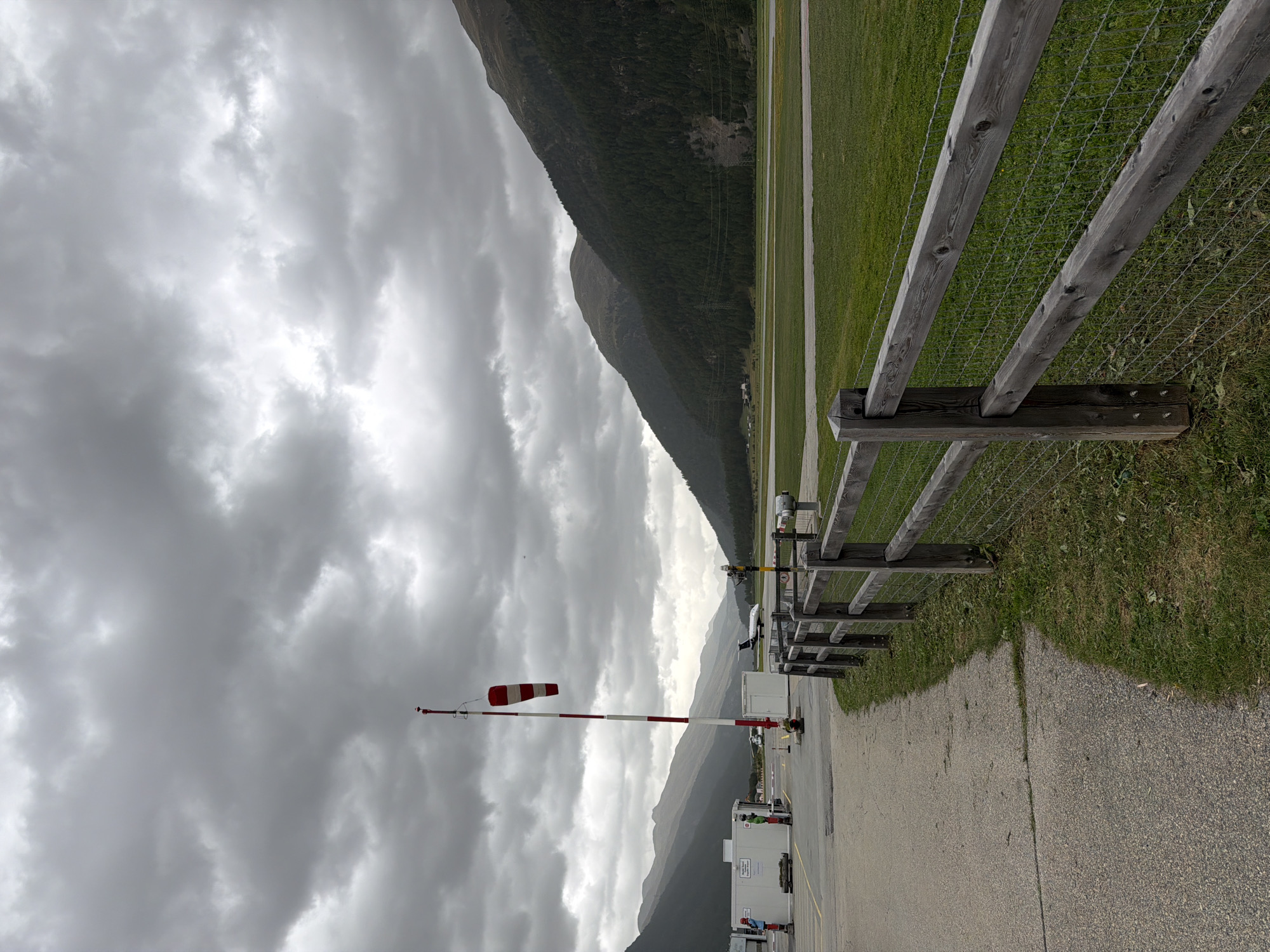 The image depicts an outdoor scene at an airport in Samedan, Switzerland, under an overcast sky. In the foreground, a weathered wooden fence with wire mesh runs horizontally across the frame, separating a paved walkway from a grassy field. A red and white striped windsock on a pole stands prominently to the left, indicating wind direction. Beyond the fence, a runway is visible, with a small jet airplane taxiing in the distance. Mountains covered in dark green trees form the backdrop, their peaks softened by the atmospheric conditions. Several white buildings, likely airport structures, are situated near the runway. Two figures, appearing to be airport personnel in uniform, are visible near one of the buildings. The overall impression is one of a functional, albeit somewhat subdued, airport environment in a mountainous region.