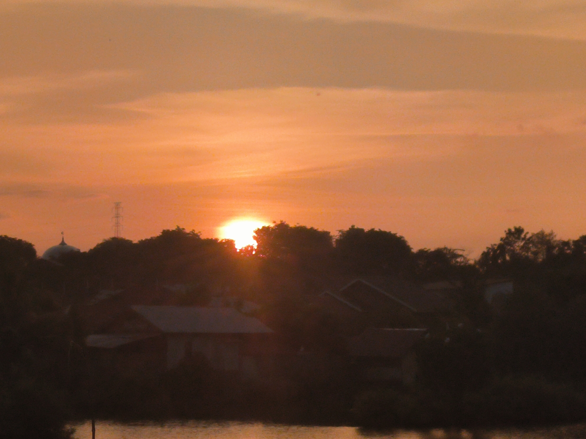 This media file captures a serene sunset or sunrise over a landscape in Lhokseumawe, Indonesia, characterized by vibrant warm tones and silhouetted foreground elements. The sky is dominated by a rich palette of orange, yellow, and soft peach hues, with diffuse clouds stretching across the upper half. The sun, a brilliant white-yellow orb, is partially visible near the center-right, just above the horizon line, casting a bright glow that obscures parts of the dark vegetation.
The foreground consists of a dark, silhouetted landscape, including dense trees and several structures. To the left, a distinct dome-shaped building, likely a mosque, stands out among the treetops. Slightly to its right, a tall, slender transmission tower or pylon is also silhouetted against the bright sky. Below these structures, other vague outlines of buildings or roofs can be discerned, blending into the dark mass of vegetation. At the very bottom of the image, a body of water, possibly a river or lake, reflects the remaining ambient light, appearing as a slightly lighter, textured strip.
The overall impression is one of tranquility, emphasizing the dramatic sky of either dawn or dusk. No specific activity or identifiable people are visible, and there is no visible text in the image. The scene conveys a sense of peaceful end or beginning to the day in a tropical setting.