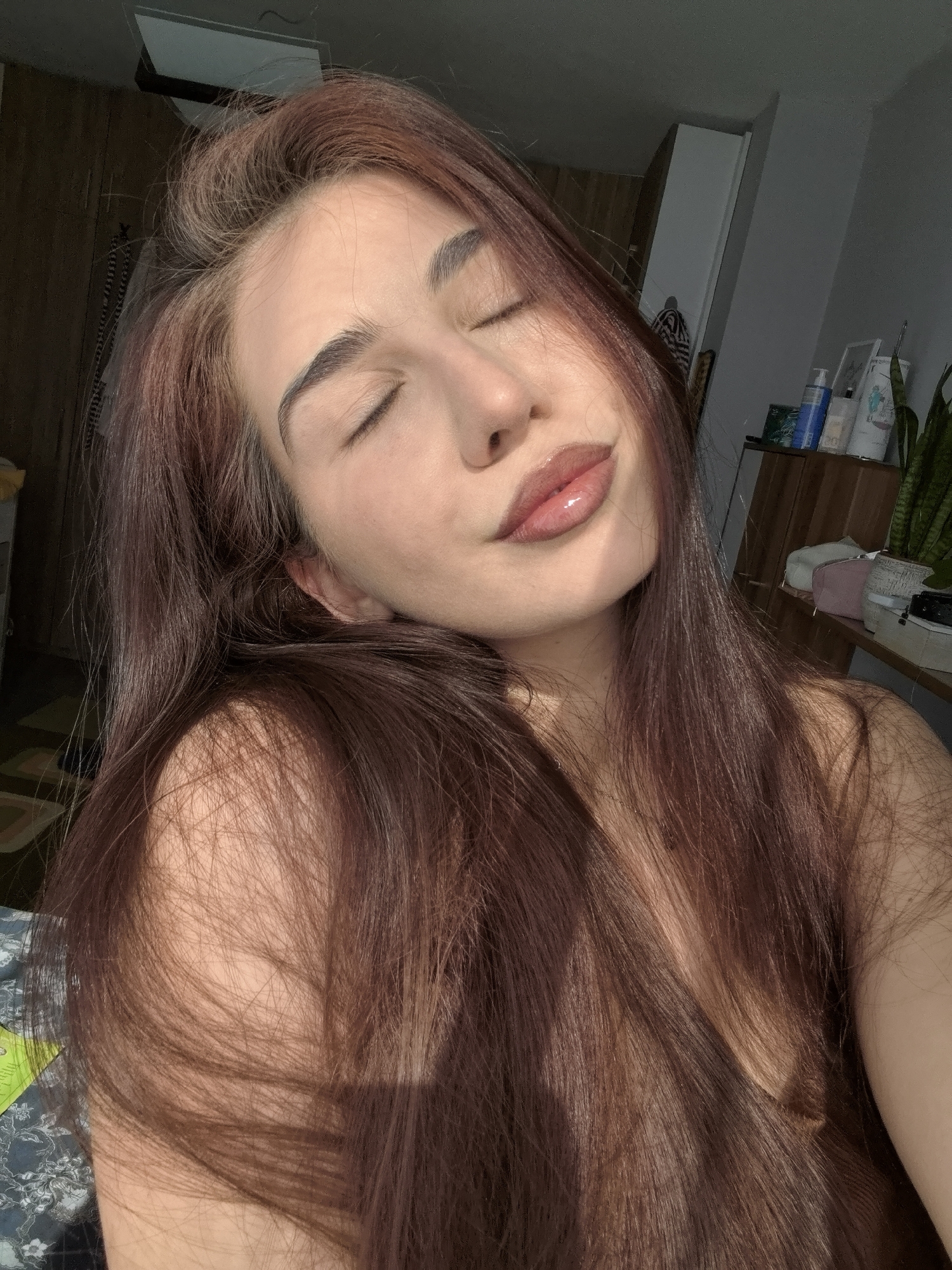 This media file presents a close-up portrait of a young woman within an indoor setting in Stara Zagora, Bulgaria. The central subject has long, flowing dark brown hair with reddish highlights, cascading over her shoulders and chest. Her eyes are closed, conveying a serene or relaxed expression, and her lips are slightly parted, adorned with a reflective gloss that catches the strong natural light. Her head is gently tilted back.
The background suggests a personal room, possibly a bedroom. To the right, a wooden dresser or shelf is visible, holding various objects including a blue bottle, several white containers, a small green houseplant (likely a snake plant), and a pink pouch. The wall behind these items is light-colored. To the left, a darker wooden piece of furniture and a glimpse of patterned fabric, possibly a pillow or blanket, can be seen.
The image is characterized by strong, direct sunlight, indicating it is daytime, possibly morning or late afternoon. This sunlight creates bright highlights on the woman's face, lips, and hair, and casts noticeable shadows, adding depth to the scene. The overall activity captured is a peaceful moment, with the woman seemingly basking in or posing with the natural light. No discernible text is visible within the image.