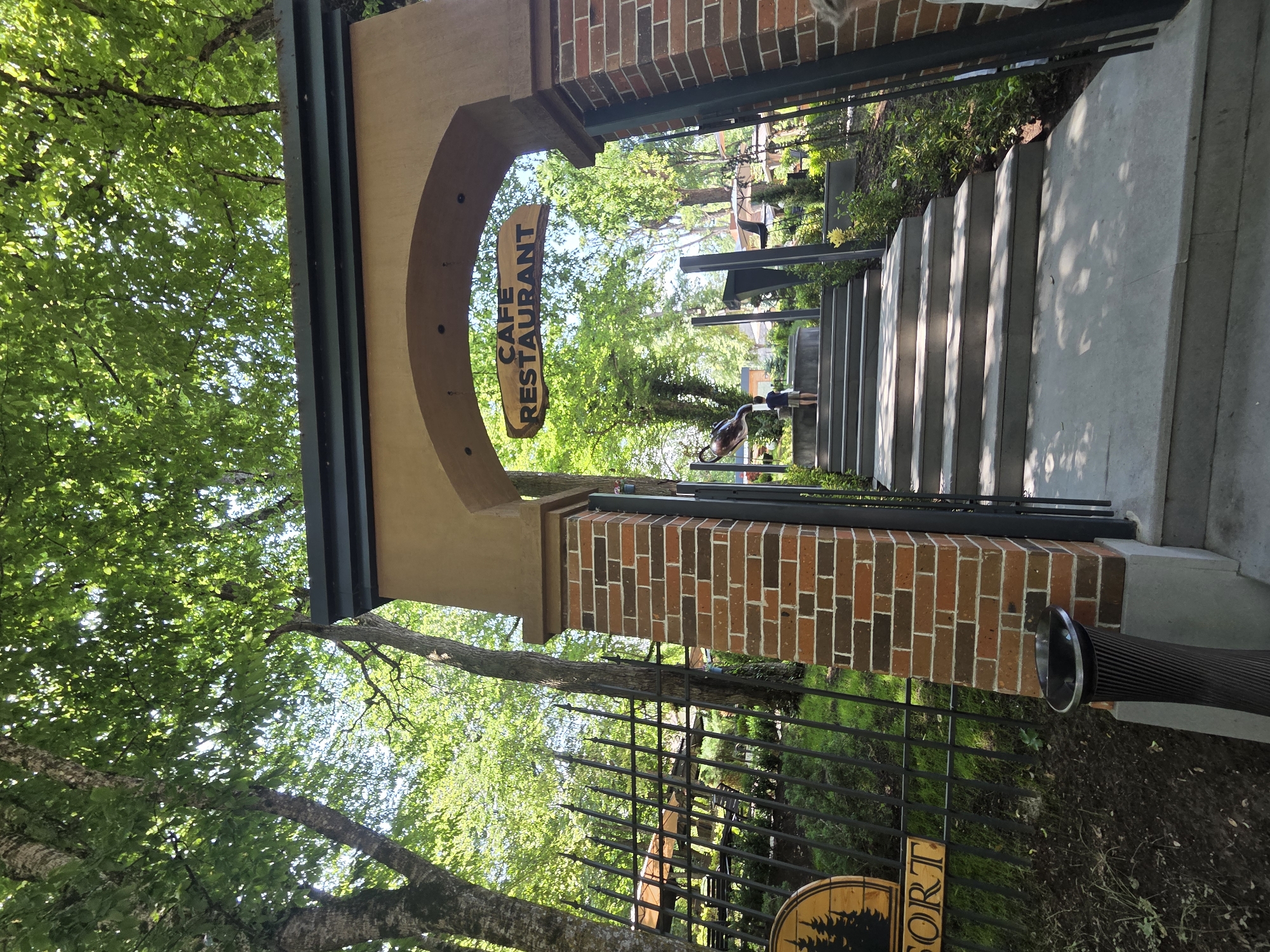 The image shows the entrance to a cafe-restaurant in Dilijan, Armenia. The entrance is an archway with brick pillars. A sign above the archway reads "CAFE RESTAURANT". A set of stairs leads up to the entrance. There are trees and greenery surrounding the entrance. A young boy is walking up the stairs. A fence with an outdoor seating area is visible to the left of the entrance.