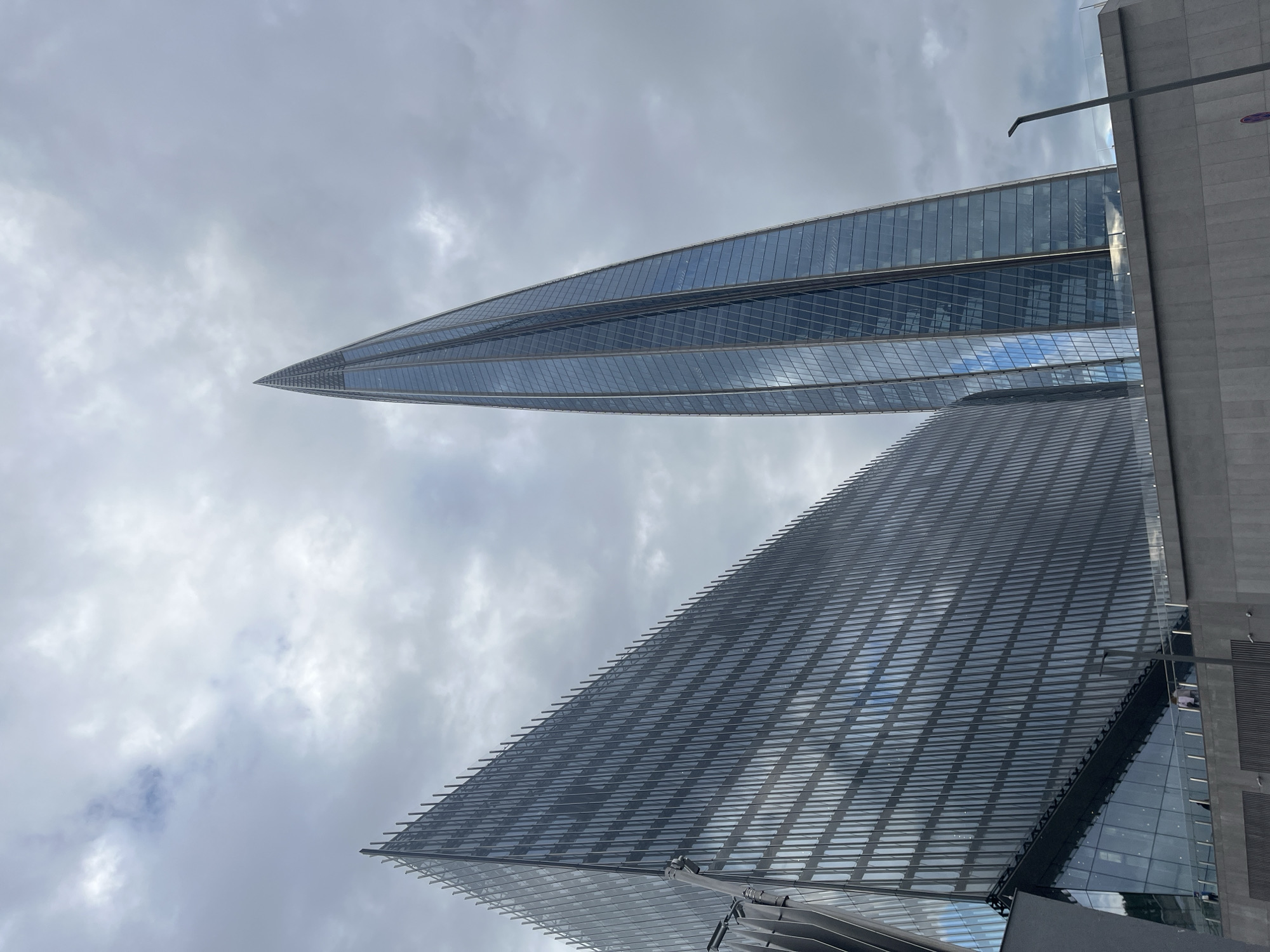 This media file captures an upward view of two modern skyscrapers with reflective glass facades against a cloudy sky. The buildings are sharply angled, with one appearing taller and more pointed, possibly tapering to a spire. The other building, in the foreground, has a more terraced or stepped design, also covered in reflective glass. The sky is overcast, with patches of lighter clouds suggesting it might be daytime but not necessarily sunny. The overall impression is one of urban architecture and modern design. There are no people or specific activities visible. The scene is dominated by the architectural forms of the buildings and the sky. Given the location context of Saint Petersburg, Russia, these could be contemporary structures within the city's developing skyline. No text is visible in the image.
