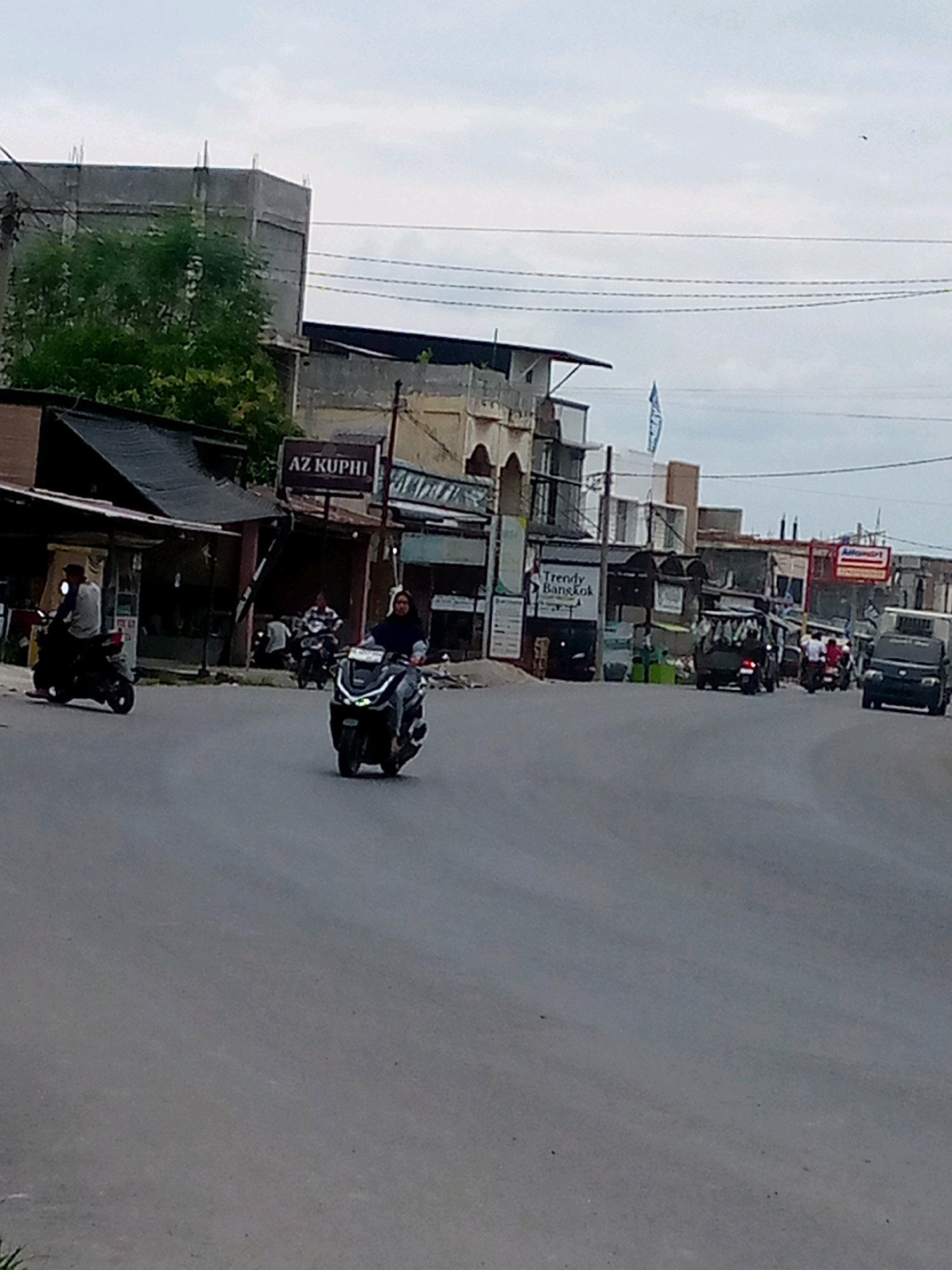 In an urban street scene, a gray asphalt road curves to the right, with a few vehicles and pedestrians visible. On the left, a person rides a black scooter. Further down the road, another individual on a black scooter is seen. In the background, buildings line the street, some appearing weathered. A sign reading "AZ KUPHI" is visible on one of the buildings. Further to the right, a van with a darkened interior is driving. A white van is parked on the side of the road. The sky is overcast.