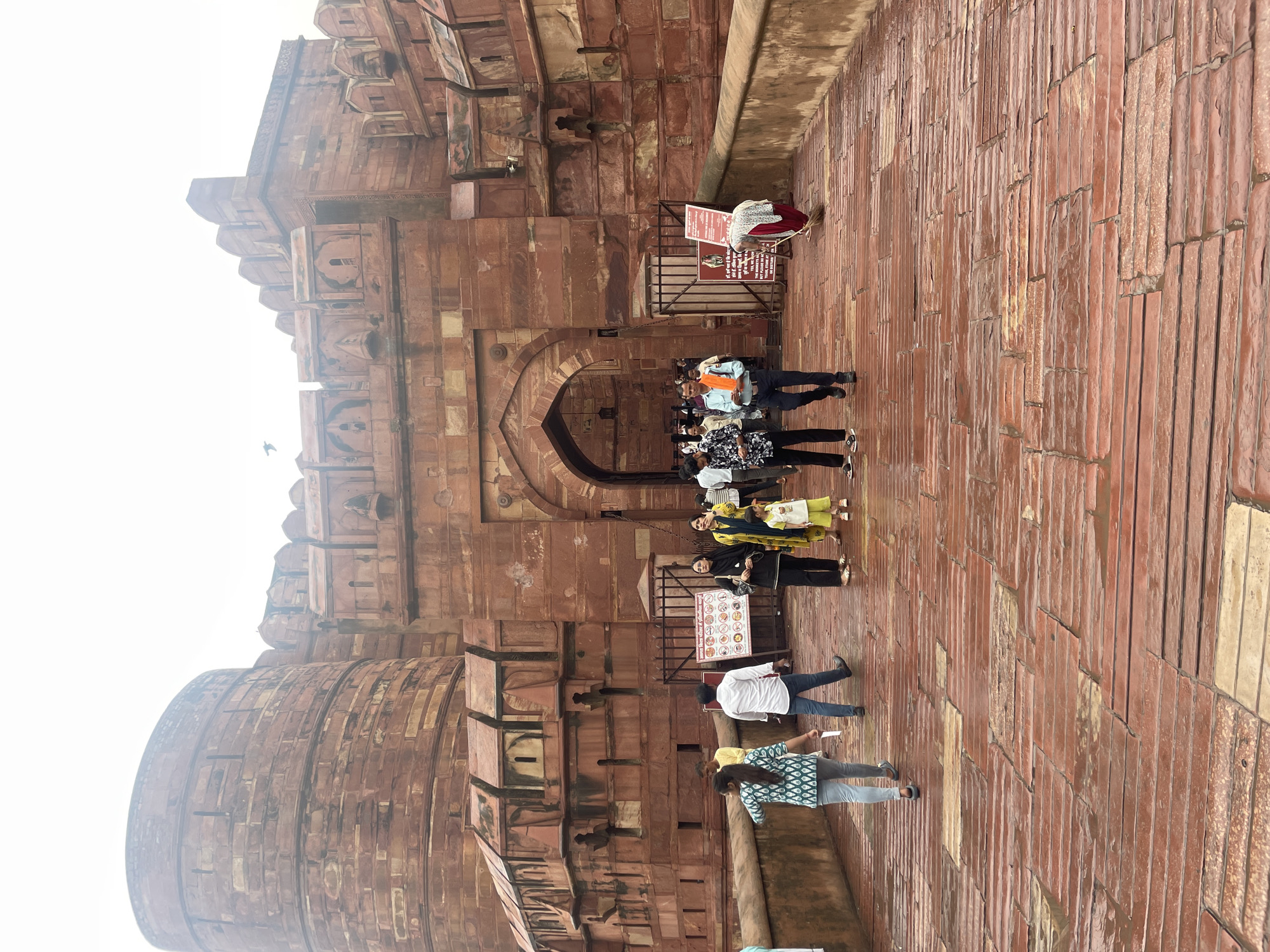 The image depicts the entrance to the Agra Fort in Agra, India. A group of tourists and locals are walking on a wet, red-tiled pathway leading to a large, arched gateway made of red sandstone. The fort's imposing architecture, characterized by massive stone walls and turrets, is visible in the background. A few people are seen conversing, while others are walking towards or away from the fort. The weather appears overcast, and the wet path suggests recent rainfall. A woman is seen sweeping the pathway. Informational signs are posted near the entrance.
