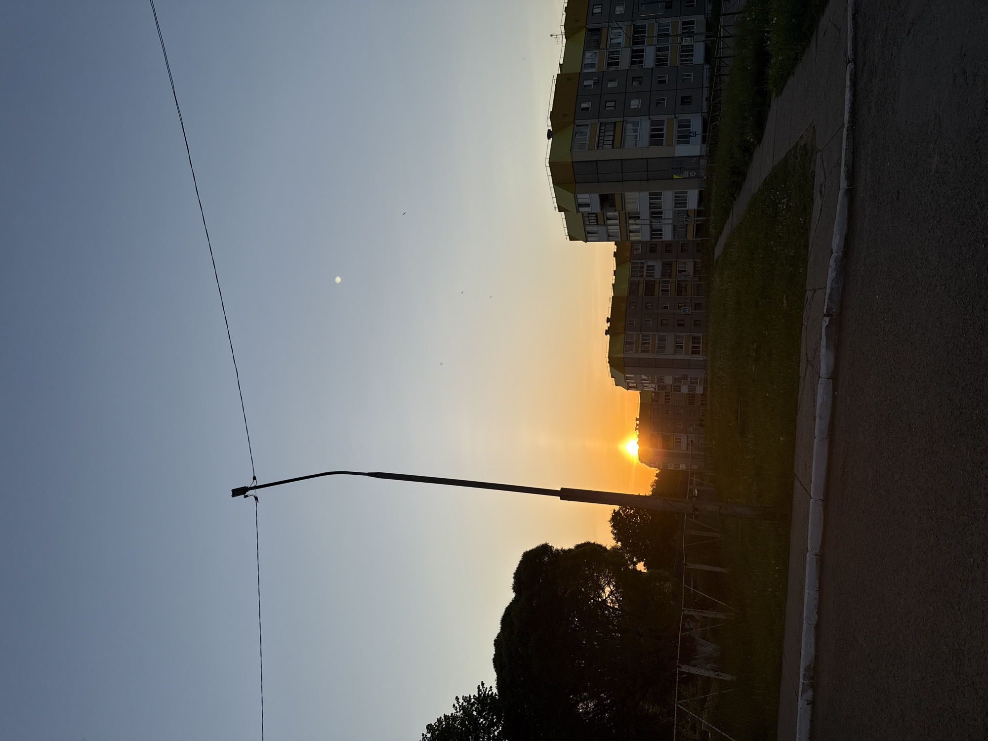 The image shows a sunset view in what appears to be a residential area. A street light pole is visible in the foreground against the backdrop of the sky transitioning from blue to orange near the horizon. A multi-story apartment building is partially visible at the top right of the frame. Trees are silhouetted in front of the setting sun. The ground shows a paved road and some grass or greenery, indicating a sidewalk or park area. Small birds are seen flying in the sky. Based on visual cues and the provided location (Марковский, Russia), this is likely a typical urban or suburban evening scene.