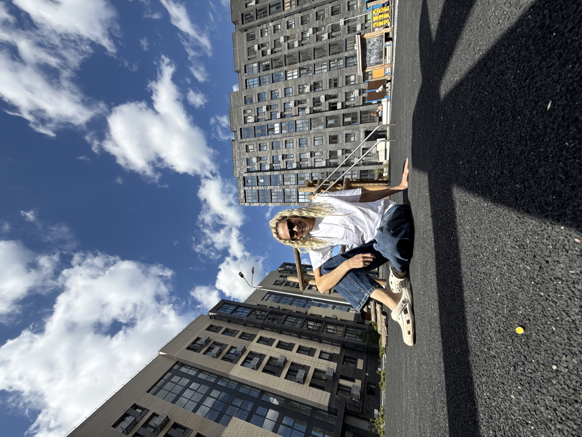 A man with long blond hair, sunglasses, a white t-shirt, and blue jeans crouches on an asphalt surface. He faces forward with his right hand touching the ground. Buildings are visible in the background. The sky is blue with scattered white clouds. A yellow object rests on the asphalt to the right of the man.