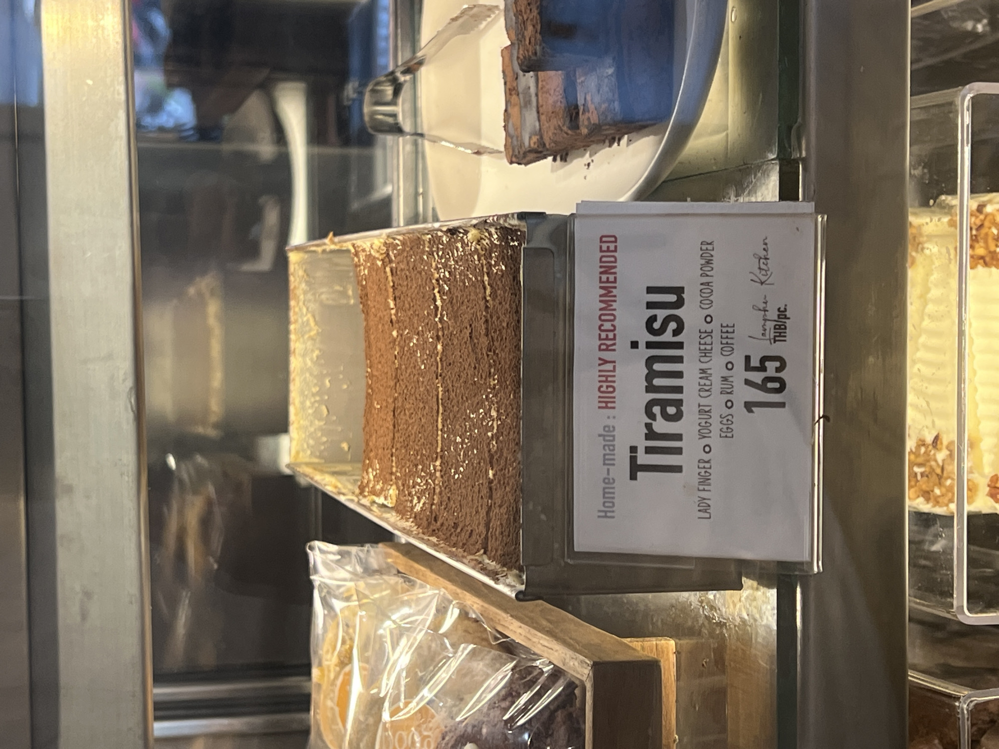 The image shows a display case containing various desserts, primarily focusing on a slice of Tiramisu. The Tiramisu, presented in a clear container, features distinct layers of coffee-soaked ladyfingers and creamy filling, topped with cocoa powder. It sits on a metallic shelf within what appears to be a refrigerated display.
To the right of the Tiramisu is a white sign with black text, oriented vertically. The sign reads: "Home-made: HIGHLY RECOMMENDED Tiramisu". Below the dessert's name, its ingredients are listed: "LADY FINGER", "YOGURT CREAM CHEESE", "COCOA POWDER", "EGGS", "RUM", and "COFFEE". At the bottom, the price "165 THB/pc." is clearly visible, along with the name "Lampha Kitchen".
Above the Tiramisu, on a higher shelf, a white plate holds what appear to be other slices of cake. Below the Tiramisu, in a wooden box or shelf, several items are visible in clear plastic packaging, suggesting other pastries or baked goods. The display case itself is made of glass and metal, reflecting blurry shapes in the background, indicating an indoor setting like a cafe or bakery in Baan Lamai, Thailand.