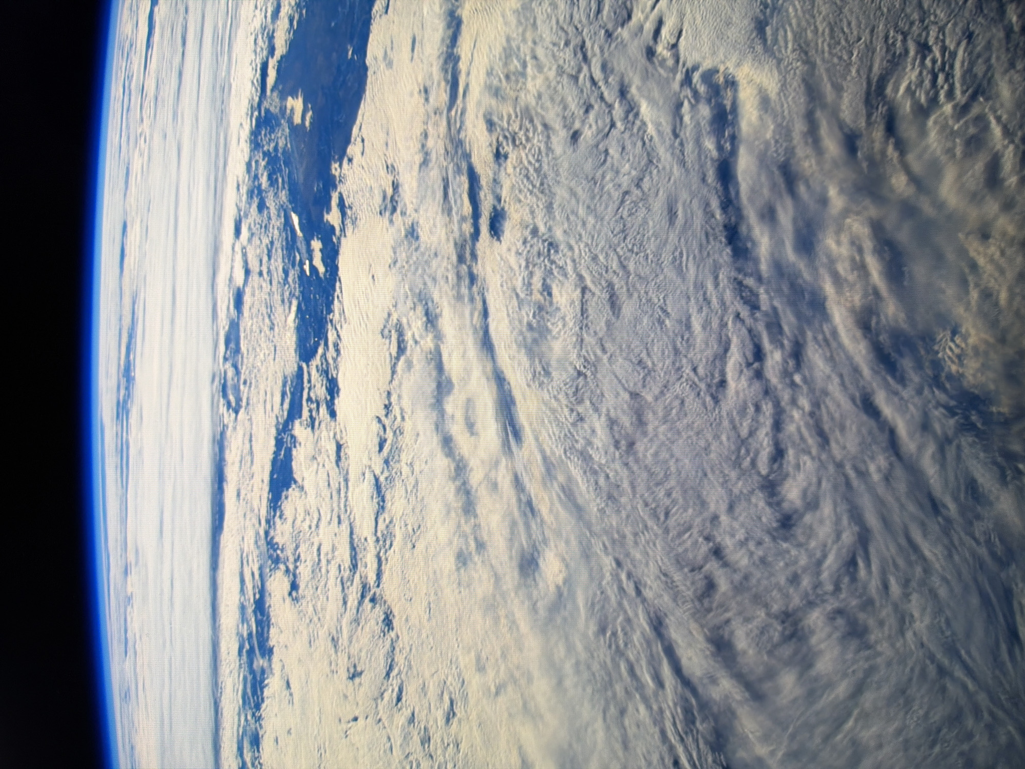 The image is a high-altitude view of the Earth's atmosphere and cloud cover, seen from space. The curvature of the planet is visible along the top edge, with a thin blue line representing the atmosphere, transitioning to the blackness of space. Below the atmosphere, vast expanses of white and grey clouds fill the frame, displaying various textures and formations. Some areas show dense cloud banks, while others reveal thinner, more wispy clouds. Sunlight illuminates the clouds from above, creating highlights and shadows that give depth to the scene. The lower part of the image shows darker blue patches, likely areas of ocean or land beneath the clouds. There are no discernible landmarks, cities, or human activity visible in this image. The scene is purely a natural, atmospheric perspective of the Earth.