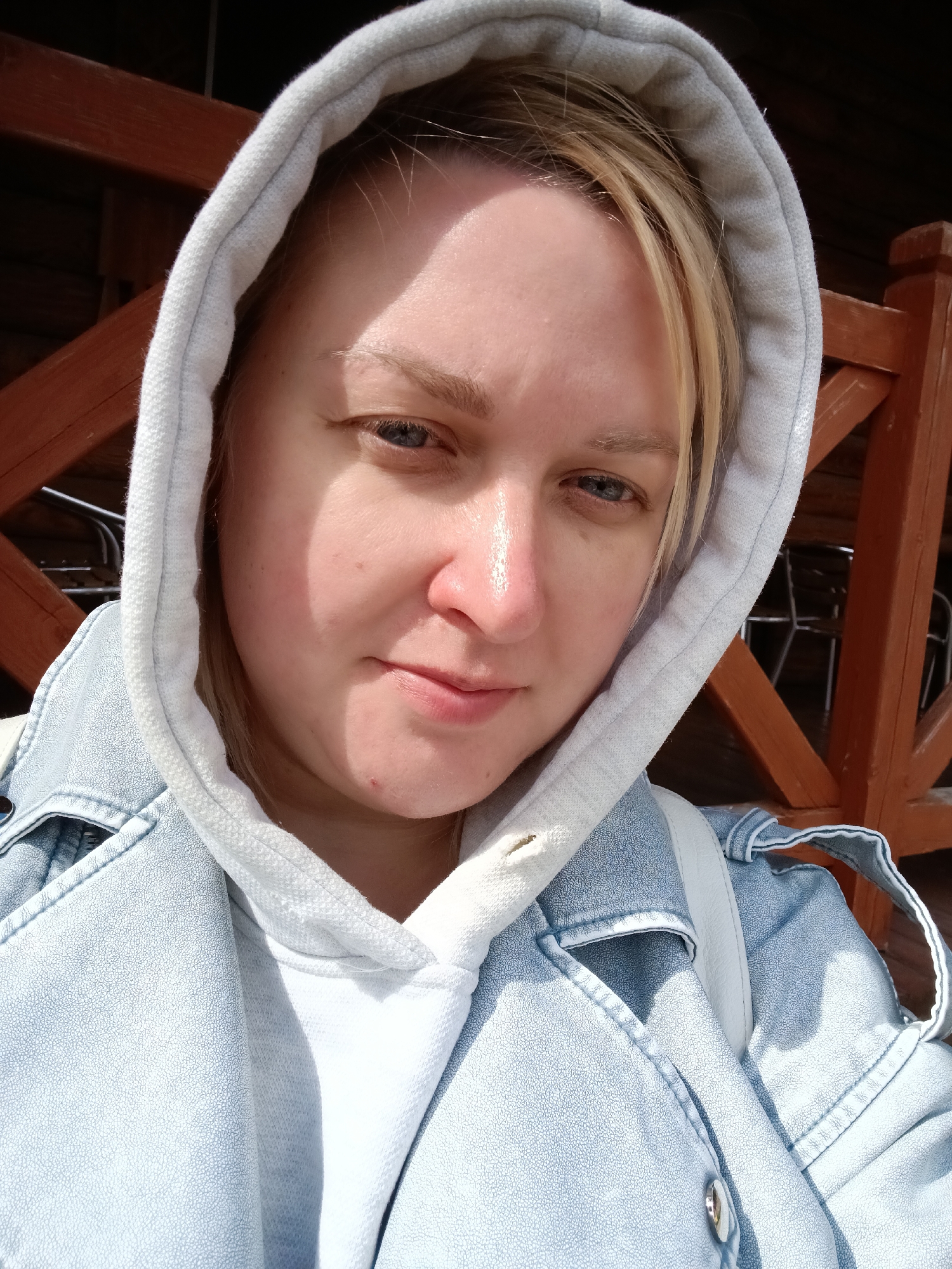A fair-skinned woman with blonde hair and blue eyes is the focus of this selfie. She is wearing a white hooded sweatshirt, with the hood up, and a light-wash denim jacket over it. The background is blurred, but appears to be a wooden structure, possibly part of a building or fence, with some dark and light brown elements. The location is specified to be in Izhevsk, Russia, but there are no immediately identifiable landmarks or features in the image to confirm this beyond the metadata. The woman is looking slightly down at the camera with a faint smile.