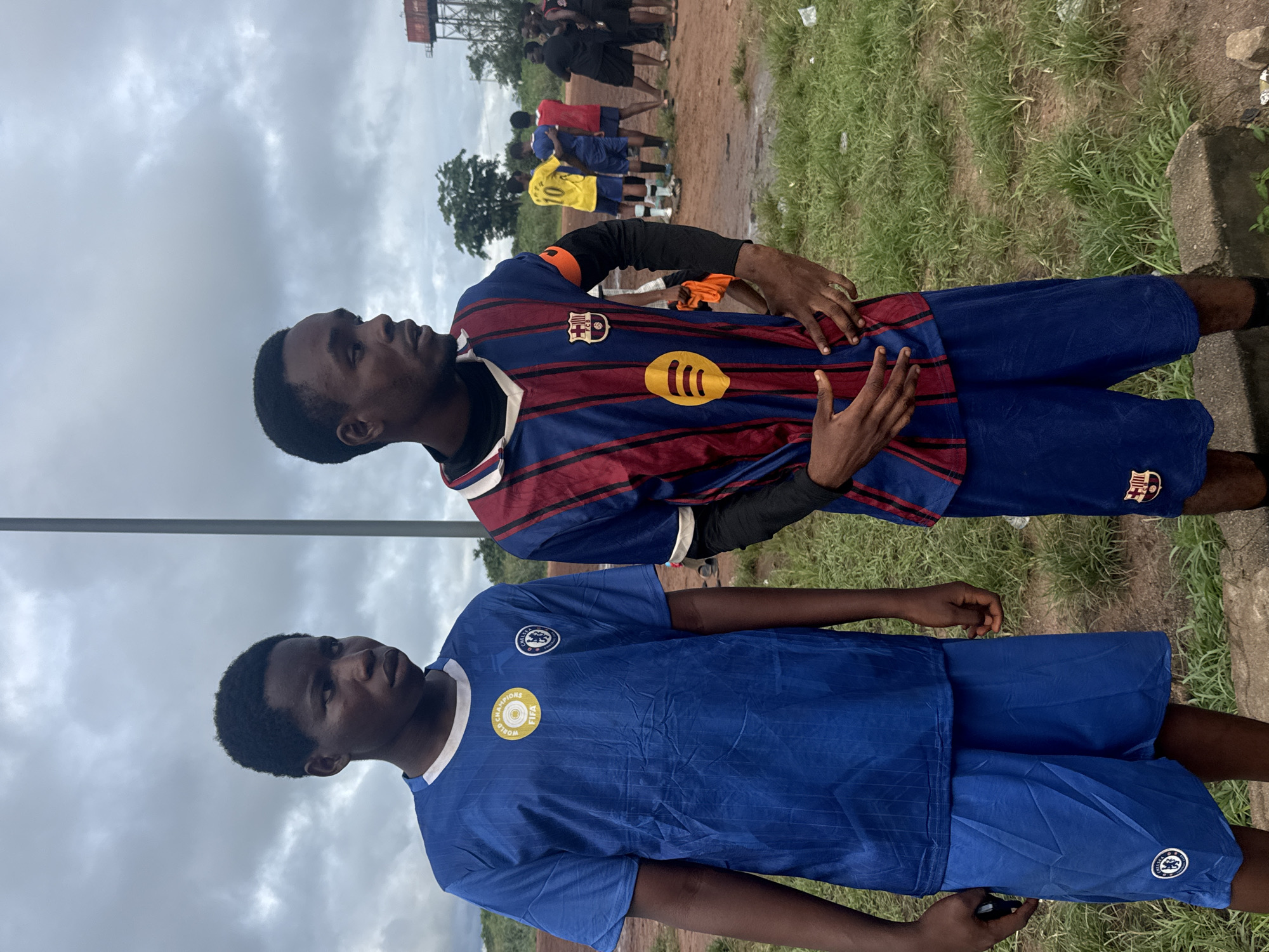 In Ihiagwa, Nigeria, two young men stand facing forward, their gazes directed slightly to the side. The man on the left wears a blue soccer jersey with white trim and a white collar, adorned with a circular FIFA badge and a club crest. He also wears matching blue shorts. The man on the right is dressed in a striped soccer jersey featuring red and blue vertical stripes, a white collar, and an orange band on his left sleeve, along with a club crest and a yellow circular logo. He is wearing blue shorts and black long-sleeved undergarments. In the background, other individuals are gathered on a dirt field, some in soccer attire, suggesting a sporting event or practice. Trees and a water tower are visible in the distance under a cloudy sky. The ground is a mixture of dirt and sparse grass, with some puddles indicating recent rainfall.