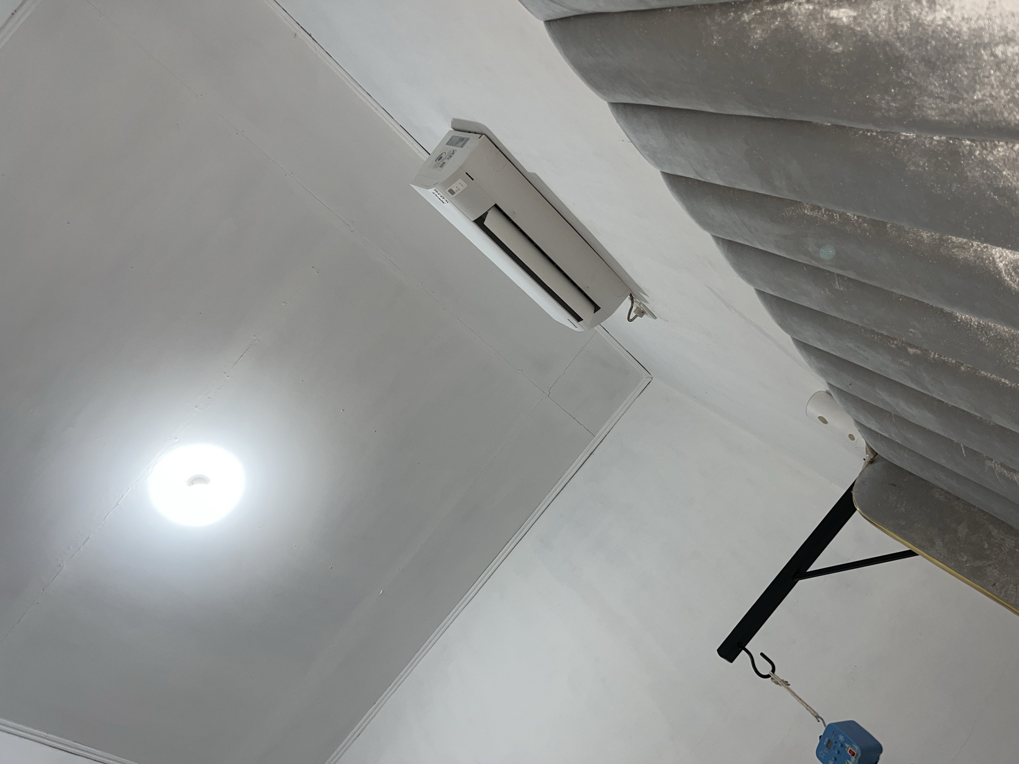 An upward-facing interior shot captures a white ceiling, a wall-mounted air conditioning unit, and a distinctive grey, textured object on the right. A bright, circular light fixture is prominently visible on the left side of the white ceiling, which displays some visible seams and minor imperfections. Centrally located, a white split-system air conditioning unit is installed, bearing the "SAMSUNG" brand name and other small text. To the right, a large grey object with a corrugated or layered texture, possibly a padded headboard or wall panel, dominates the frame. A black metal bracket extends from the right, supporting a rope with a hook from which a small blue device, resembling a digital weighing scale with a screen and buttons, is suspended. The scene depicts a static indoor environment, likely a room in Lhokseumawe, Indonesia. No specific activity or individuals are present.