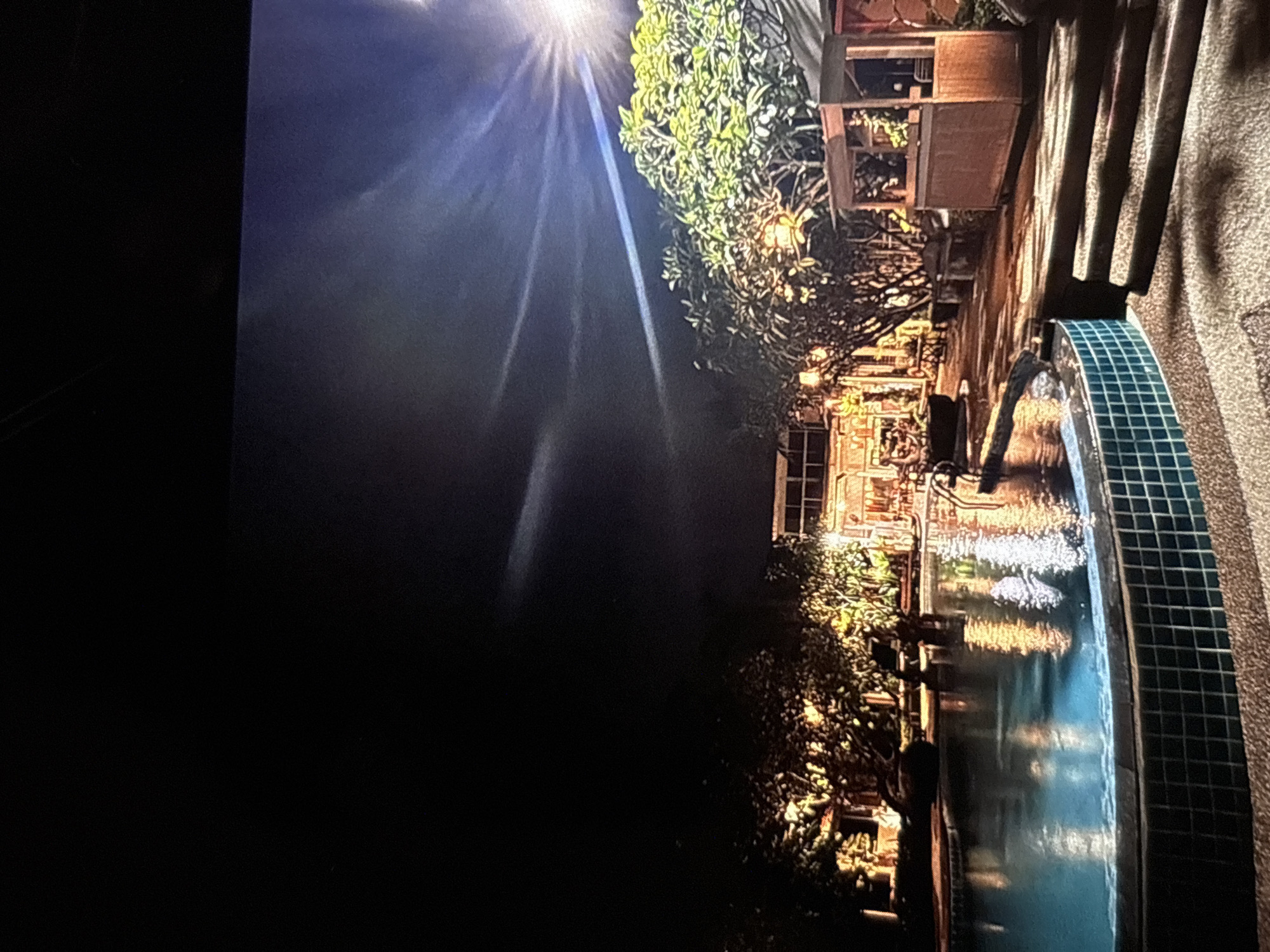 The image depicts an outdoor swimming pool area at night in Chalong, Thailand. The pool itself is illuminated by lights that reflect off its surface, creating a vibrant blue hue. The pool is bordered by mosaic tiles and a sandy pathway. Surrounding the pool are lush green trees, some of which are also lit, casting warm yellow light. In the background, there is a building with multiple windows, appearing to be a hotel or resort, also illuminated at night. A small wooden structure, possibly a kiosk or bar, is visible to the right. A bright light source, likely a floodlight, is positioned high in the sky, casting strong beams that streak across the dark sky. There are no visible people in the image.