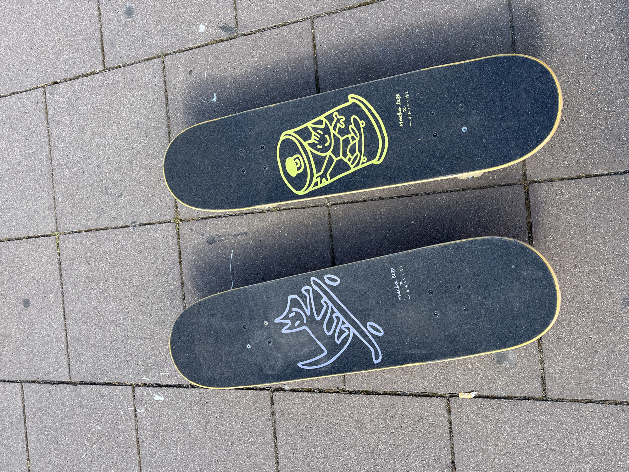 Two skateboards rest on a paved surface of gray rectangular bricks. The top skateboard features a graphic of a yellow can with a cartoon figure inside. The bottom skateboard has a graphic of a white abstract design. Both skateboards are black and have the words "Macha Life x Mariscal" printed on them. The location is likely Barcelona, Spain due to the skateboards being a collaboration between the Macha Life brand and artist Javier Mariscal, who is based in Barcelona.