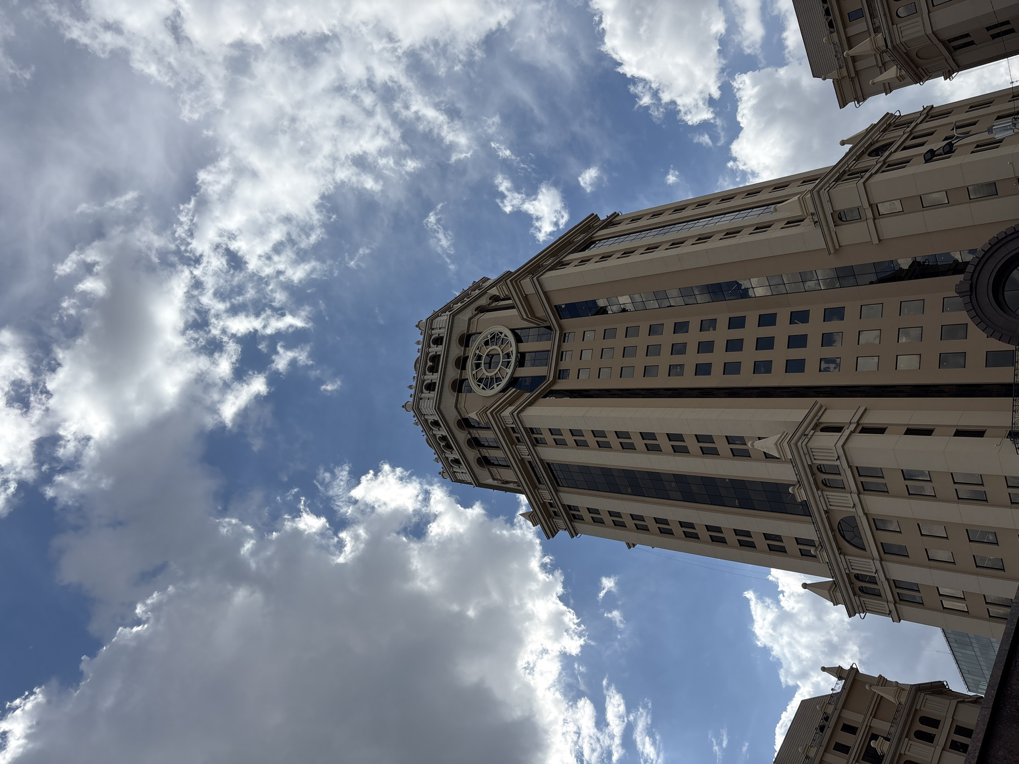 A low-angle, full-shot photograph captures the imposing facade of the building known as the Stalin-era skyscraper on Kotelnicheskaya Embankment in Moscow, Russia. The building, characterized by its beige facade and numerous dark-tinted windows, dominates the frame, stretching towards a sky filled with dramatic, fluffy white clouds against a clear blue backdrop. A large, ornate clock face is visible near the top of the skyscraper, a prominent architectural feature. The upper portion of the building is adorned with decorative architectural elements, including what appear to be crenellations and arched windows. To the left and right edges of the frame, portions of other, similar Stalin-era buildings are visible, suggesting a neighborhood of grand architecture. The overall impression is one of architectural grandeur set against a dynamic sky.