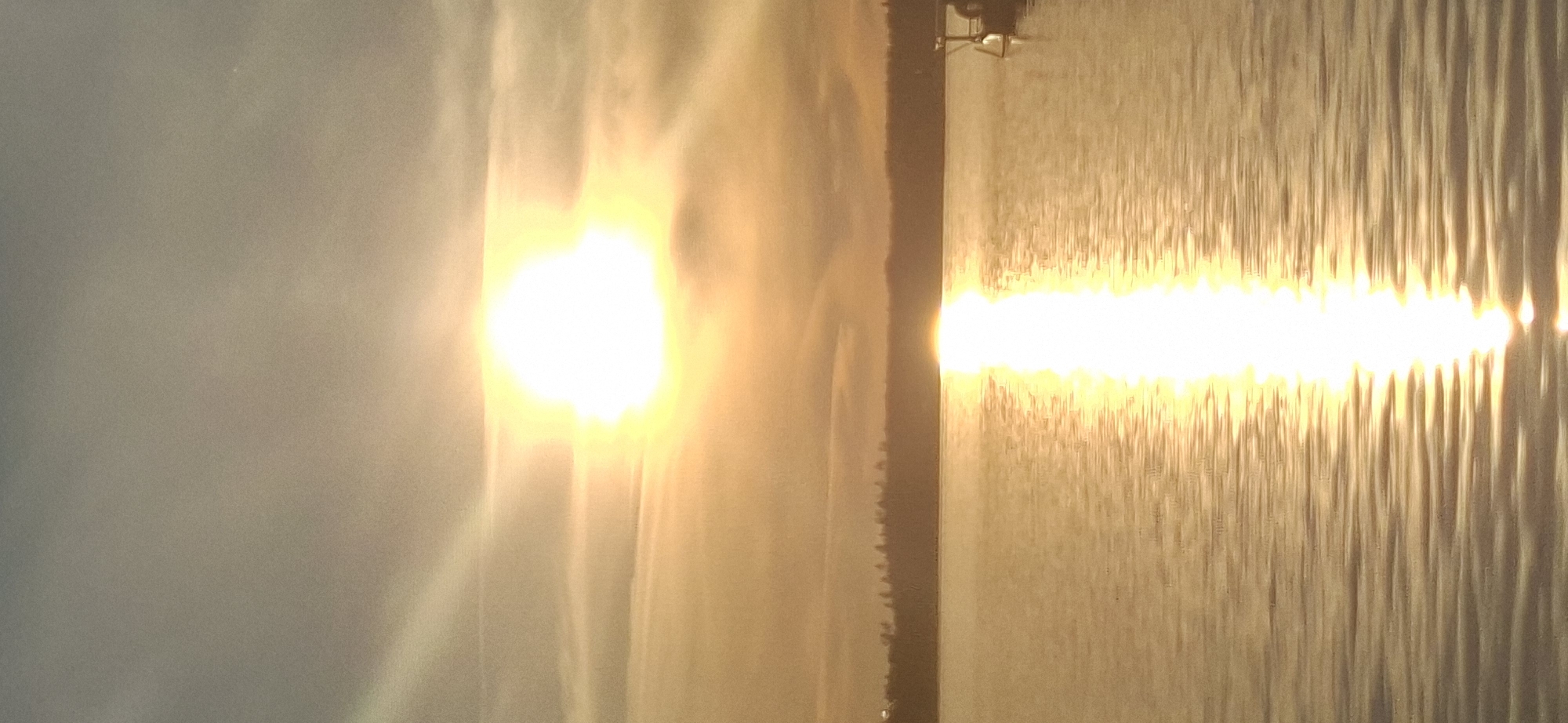 The image shows a sunset over a body of water, likely a lake or a bay, with a silhouetted treeline in the background. The sun is setting on the horizon, casting a warm, golden glow across the sky and water. A bright reflection of the sun shimmers on the surface of the water, creating a path of light. There is a small dock or pier visible on the right side of the image with a small structure. The location is identified as Salpakangas, Finland, suggesting this is a view of the natural landscape surrounding the city. The overall mood is peaceful and serene, characteristic of a sunset scene.
