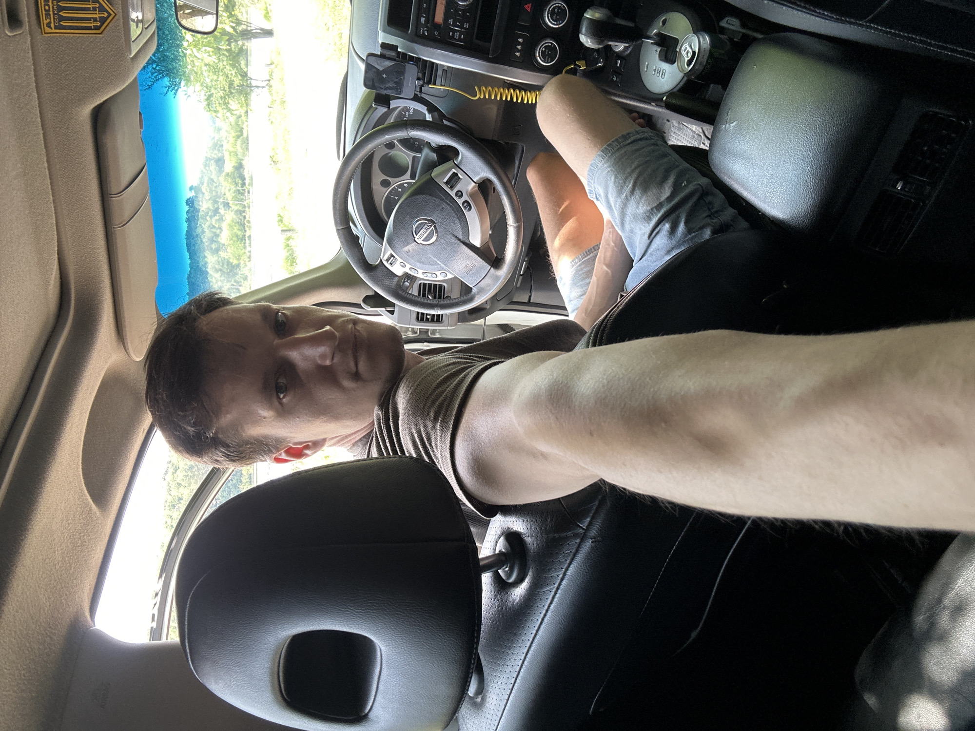 The image shows a man sitting in the driver's seat of a car. He is wearing a brown t-shirt and blue shorts. The car appears to be parked outdoors, with greenery visible through the windows. There is a phone mounted on the dashboard. A can of beverage is visible in the cup holder. The man is looking towards the camera, and his right arm is extended, holding the camera. The car's interior, including the steering wheel, dashboard, and center console, is visible. A patch with the "AZOV" logo and text "ССОВ КИЇВ" is affixed to the sun visor. The overall impression is a candid snapshot of a person in a vehicle on a sunny day. The location is confirmed as Makiv, Ukraine, by the "AZOV" insignia.