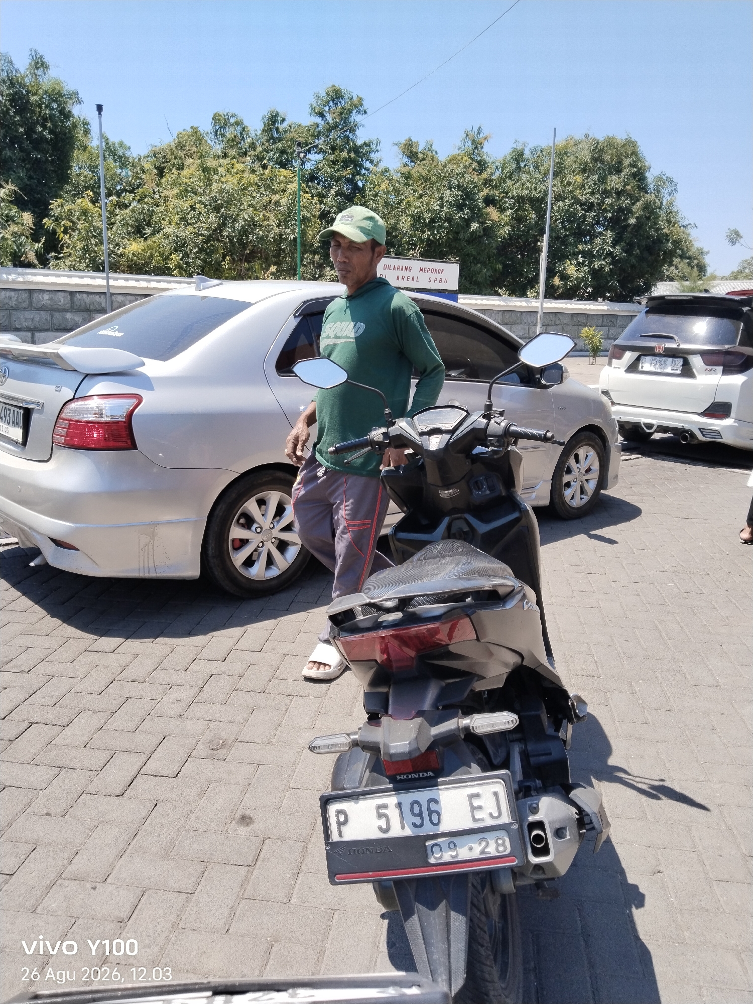 In Situbondo, Indonesia, under a clear blue sky, a man wearing a green cap and green t-shirt stands next to a silver sedan and a black motorcycle. He is holding the handlebars of the motorcycle, appearing to be either mounting or dismounting it. The man is dressed in casual attire, with striped pants and white sandals. The sedan is parked behind him, and a white SUV is visible in the background. A sign with Indonesian text "DILARANG MEROKOK DI AREAL SPBU" (No Smoking in the Gas Station Area) is mounted on a pole, suggesting the location might be near a gas station. The ground is paved with interlocking bricks. The timestamp "vivo Y100 26 Agu 2026, 12.03" is overlaid on the bottom left corner of the image.