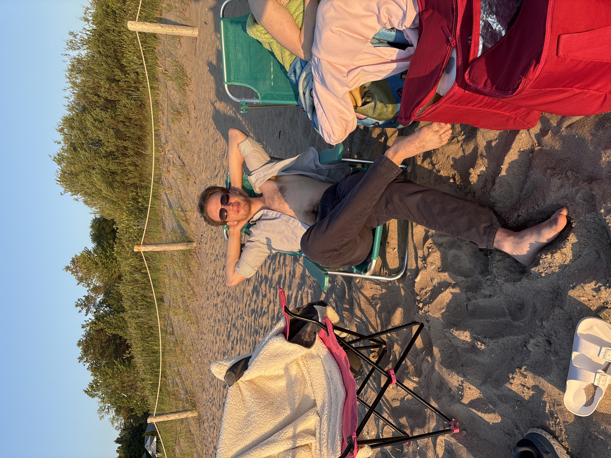 This image depicts a sunny day on the sandy shores of Wasaga Beach, Canada. In the foreground, a man in his 20s with light brown hair and sunglasses is relaxing in a green beach chair, his arms behind his head. He is wearing a light-colored, unbuttoned shirt, revealing his chest, and dark pants. To his left, a pink beach chair holds a fluffy white blanket. To his right, another green beach chair is occupied by a person whose legs and torso are visible, covered by a pink shirt and a green towel. Several items are scattered on the sand: a pair of dark sneakers, white sandals, and a large red cooler bag. A rope barrier with wooden posts separates the beach from vegetation and trees in the background. The lighting suggests it is late afternoon or early evening.