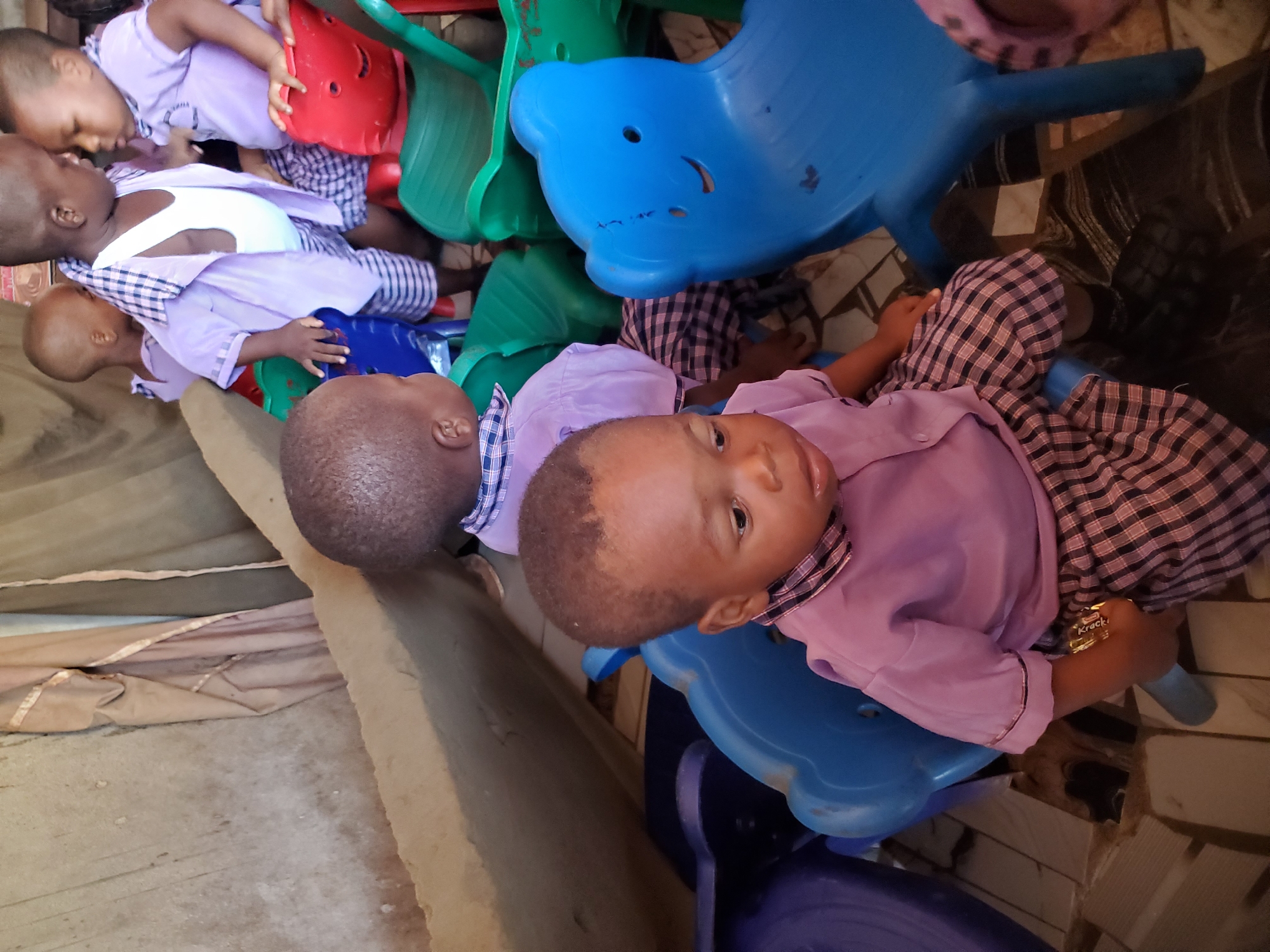 The image captures a group of young children, likely toddlers or preschoolers, in what appears to be an informal indoor setting, possibly a classroom or play area in Ukwu-Abwa, Nigeria. There are at least five children visible, all with dark skin and short hair. They are uniformly dressed in light purple long-sleeved shirts with plaid collars and matching plaid shorts or skirts.
The children are interacting with and resting on various brightly colored plastic chairs and rockers. Dominant colors of the plastic furniture include blue, green, and red. One large blue plastic object in the center-top resembles an animal-shaped rocker, possibly a whale or dolphin.
In the foreground, one child, dressed in purple and plaid, is sitting or lying on a blue plastic chair, looking directly at the camera with a neutral expression. Another child to the left of the foreground child is lying down, partially obscured. Further to the left, more children are seen, with one lying down on another blue plastic chair/rocker, and two others in the upper left corner, one seemingly interacting with another child while sitting on a red plastic chair. One child's hand holds a snack wrapper with the word "Krack" clearly visible.
The flooring in the scene varies; on the left, it appears to be a light-colored, possibly dirt or concrete surface, partially covered by a beige fabric or mat. On the right, the floor transitions to patterned tiles. The lighting suggests it is daytime. The overall impression is one of children at rest or engaged in quiet play in a simple, functional environment.